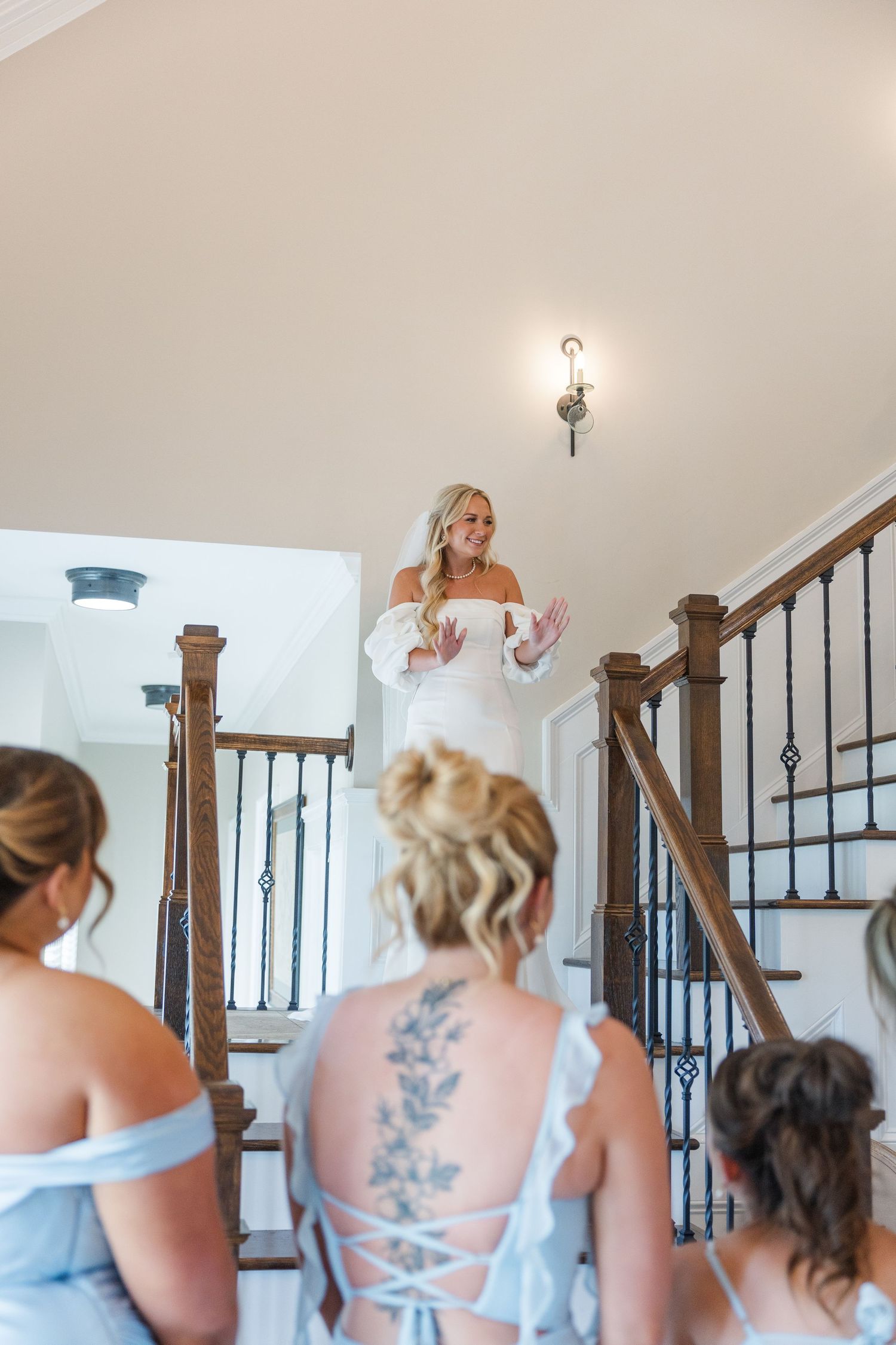 Blonde bride in white gown descending staircase revealed to bridesmaids in light blue dresses.