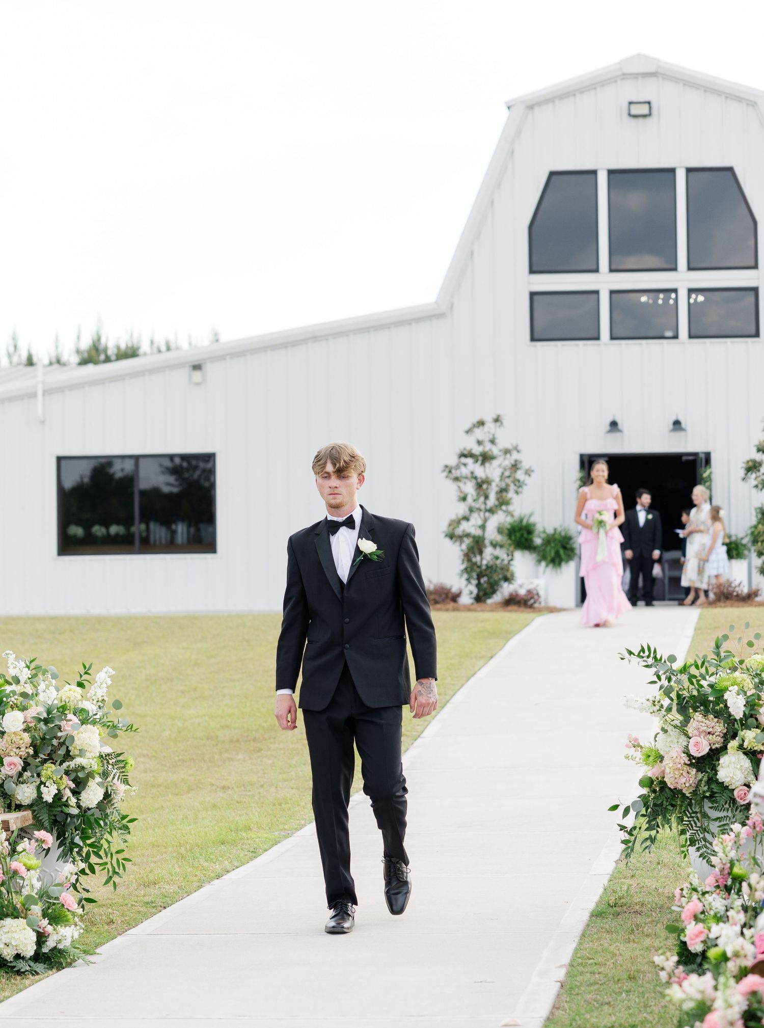 Young groomsman in black tuxedo walks solo down flower-lined wedding aisle toward white barn venue.