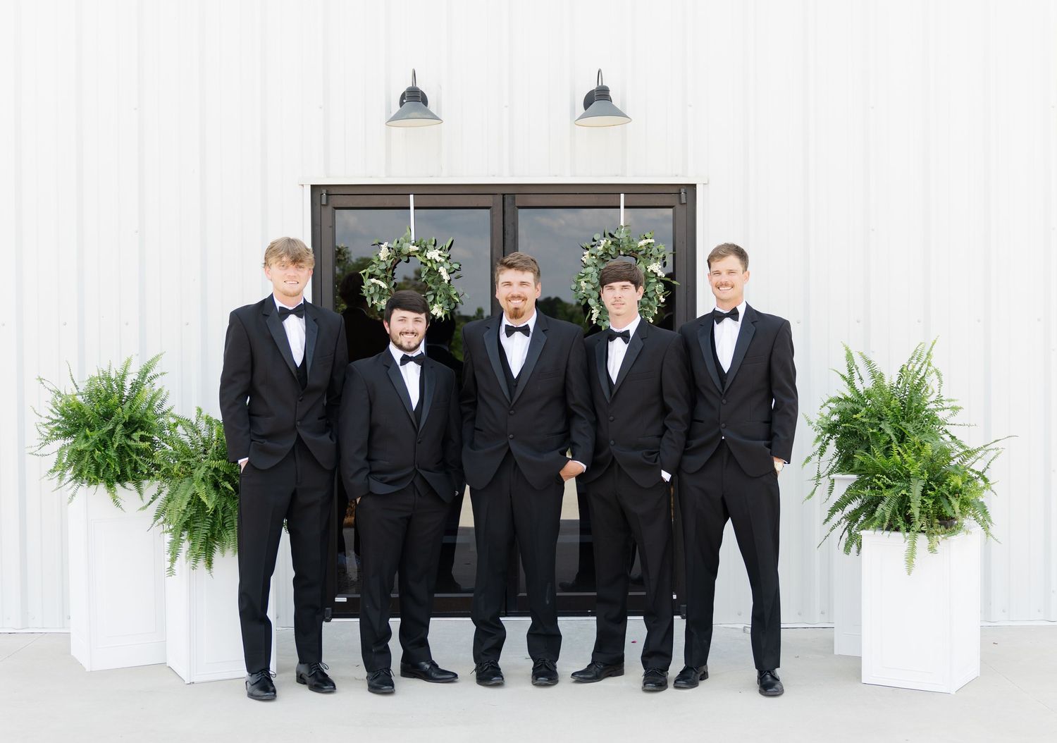 Five groomsmen in black tuxedos with bow ties posing in front of white barn doors with greenery wreaths.