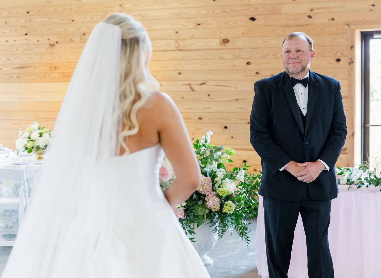 Bride in white gown and veil shares emotional first look moment with father in black tuxedo.