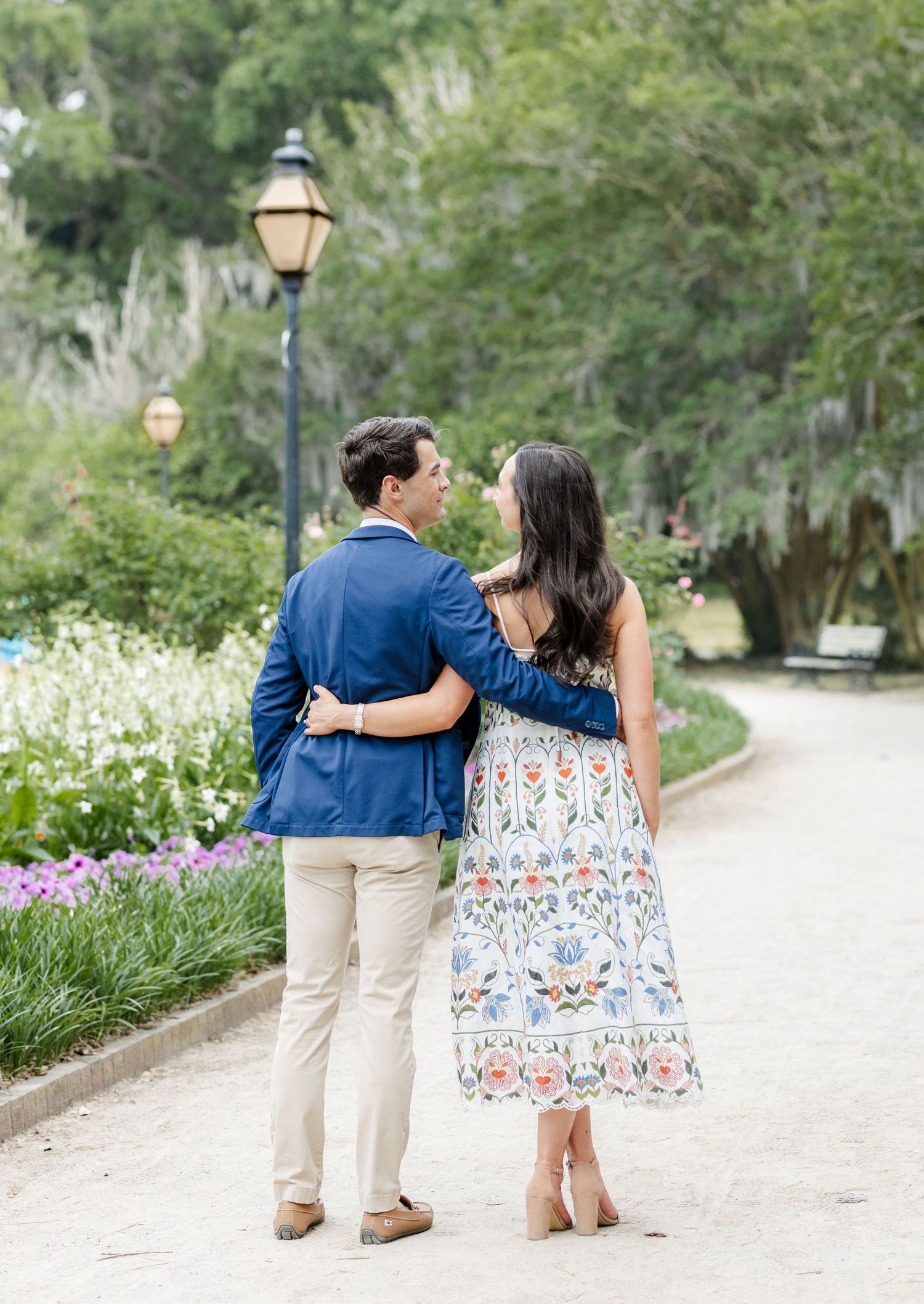 Couple walks arm in arm down a scenic garden path, man in navy blazer and woman in floral midi dress.