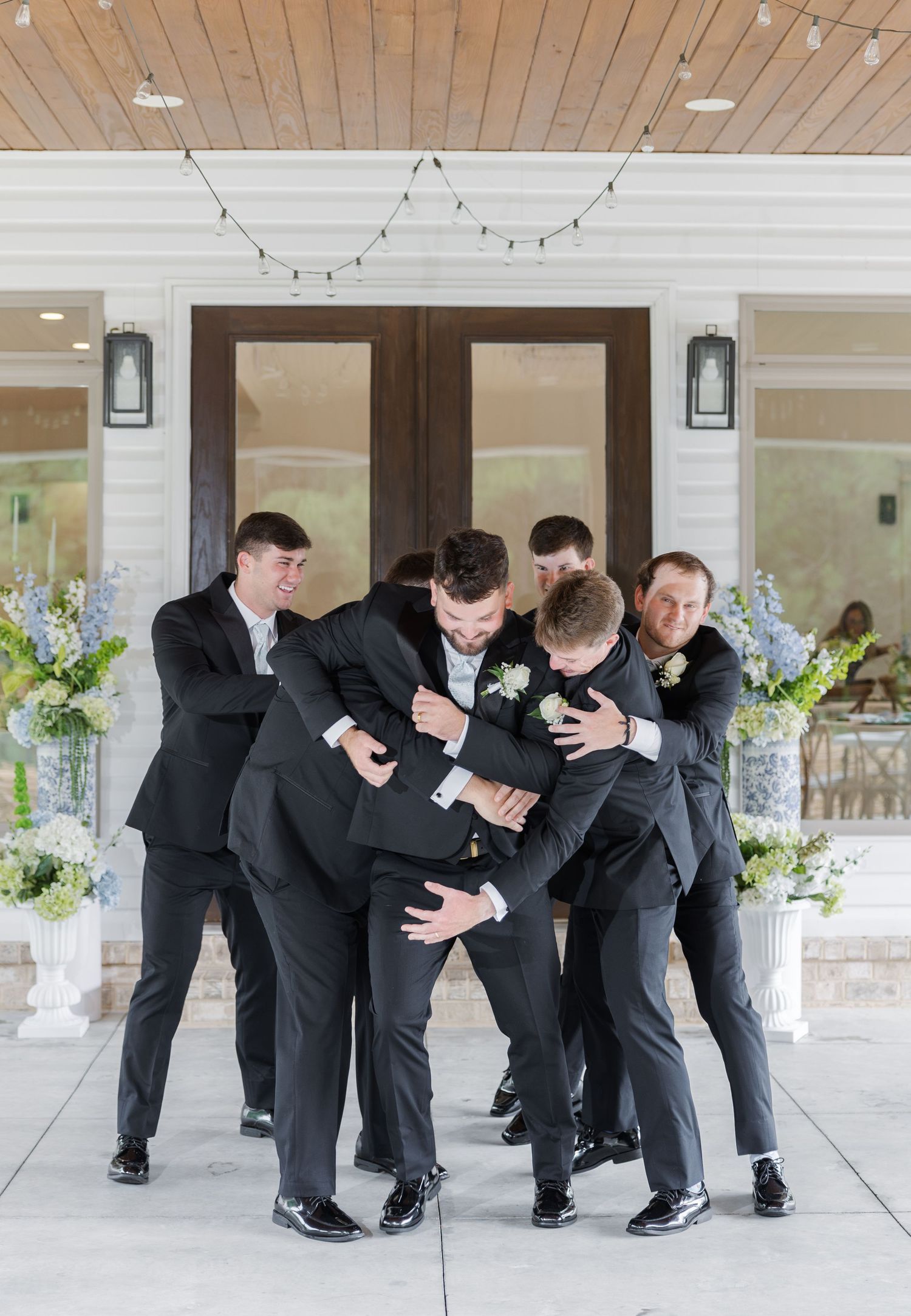 Groomsmen playfully pile on the groom in black suits outside wedding venue entrance.