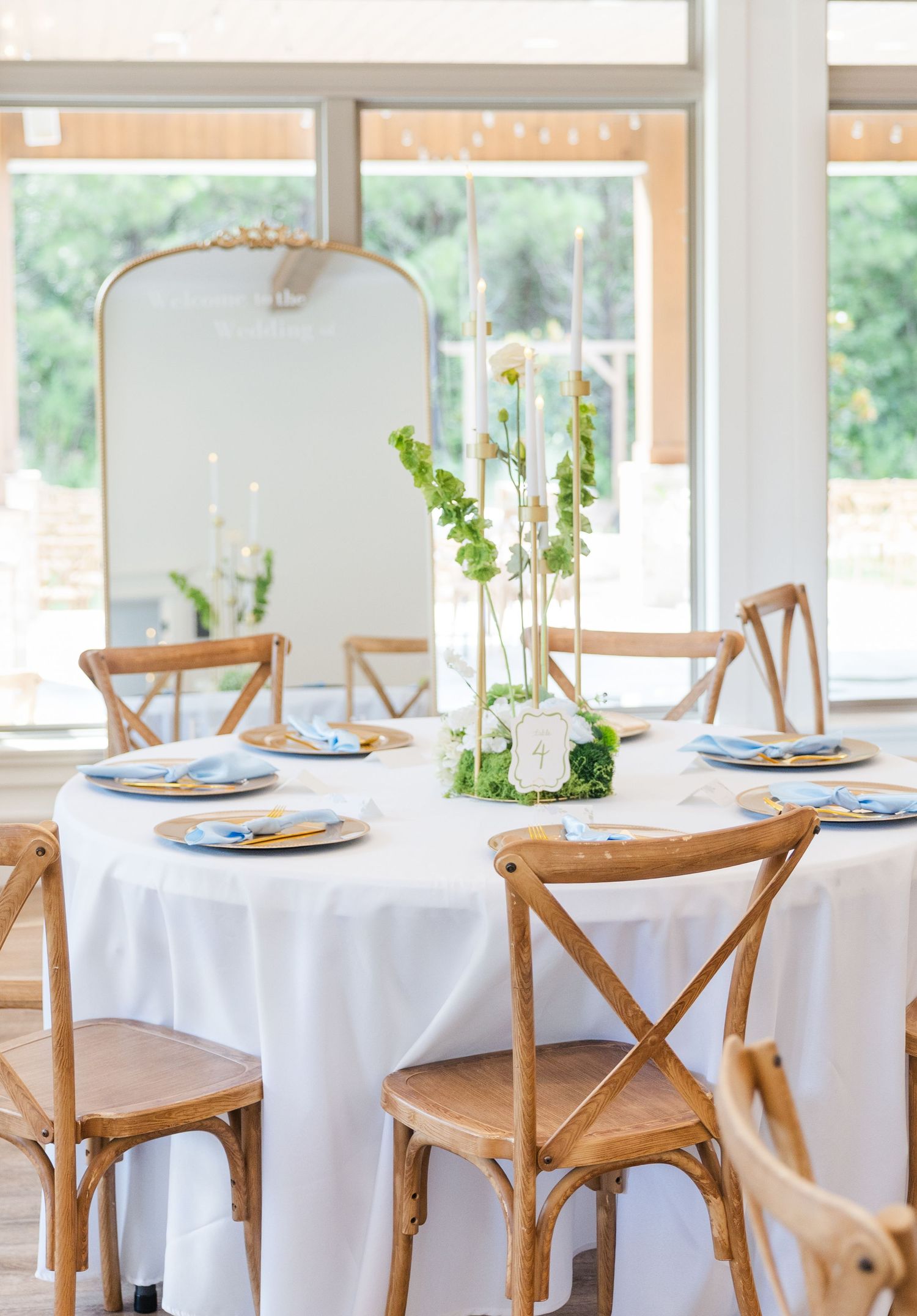 Round wedding reception table with white linen, wooden cross-back chairs, and tall green floral centerpiece.