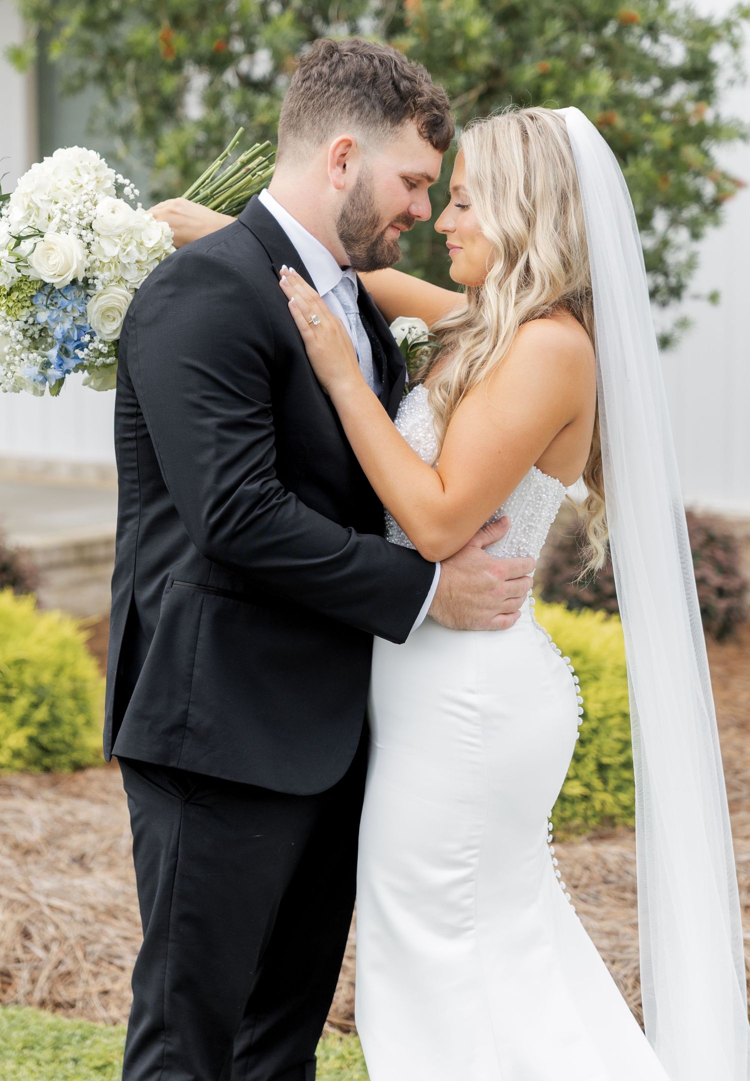 Bride and groom sharing romantic forehead touch moment outdoors holding white and blue floral bouquet.