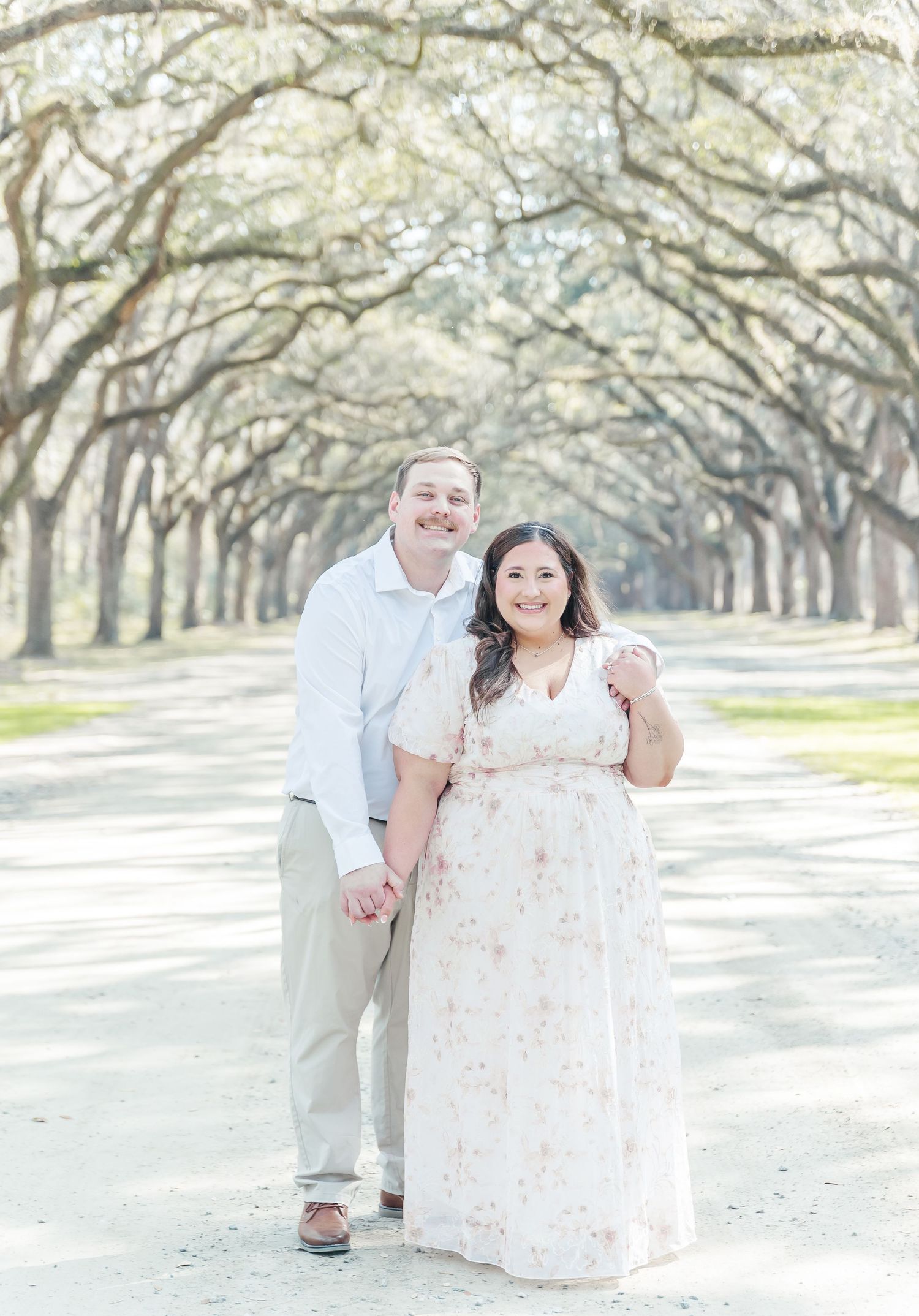 Smiling couple poses on a majestic oak-lined path during their engagement photography session.