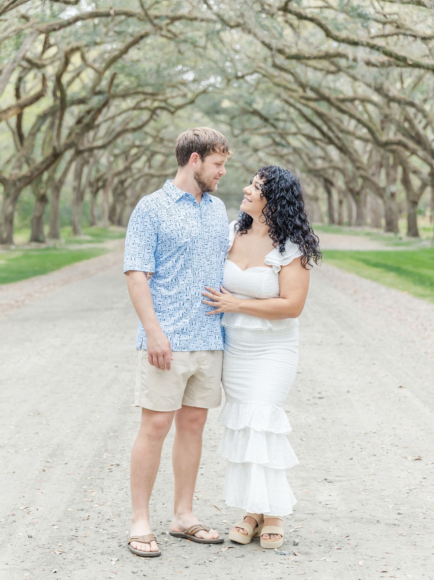 Couple in light summer attire share a romantic moment on a tree-lined path.