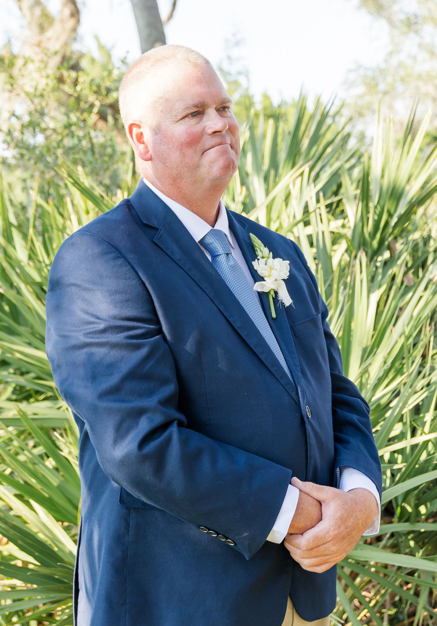 Groomsman in navy suit with white boutonniere stands in front of tropical foliage.