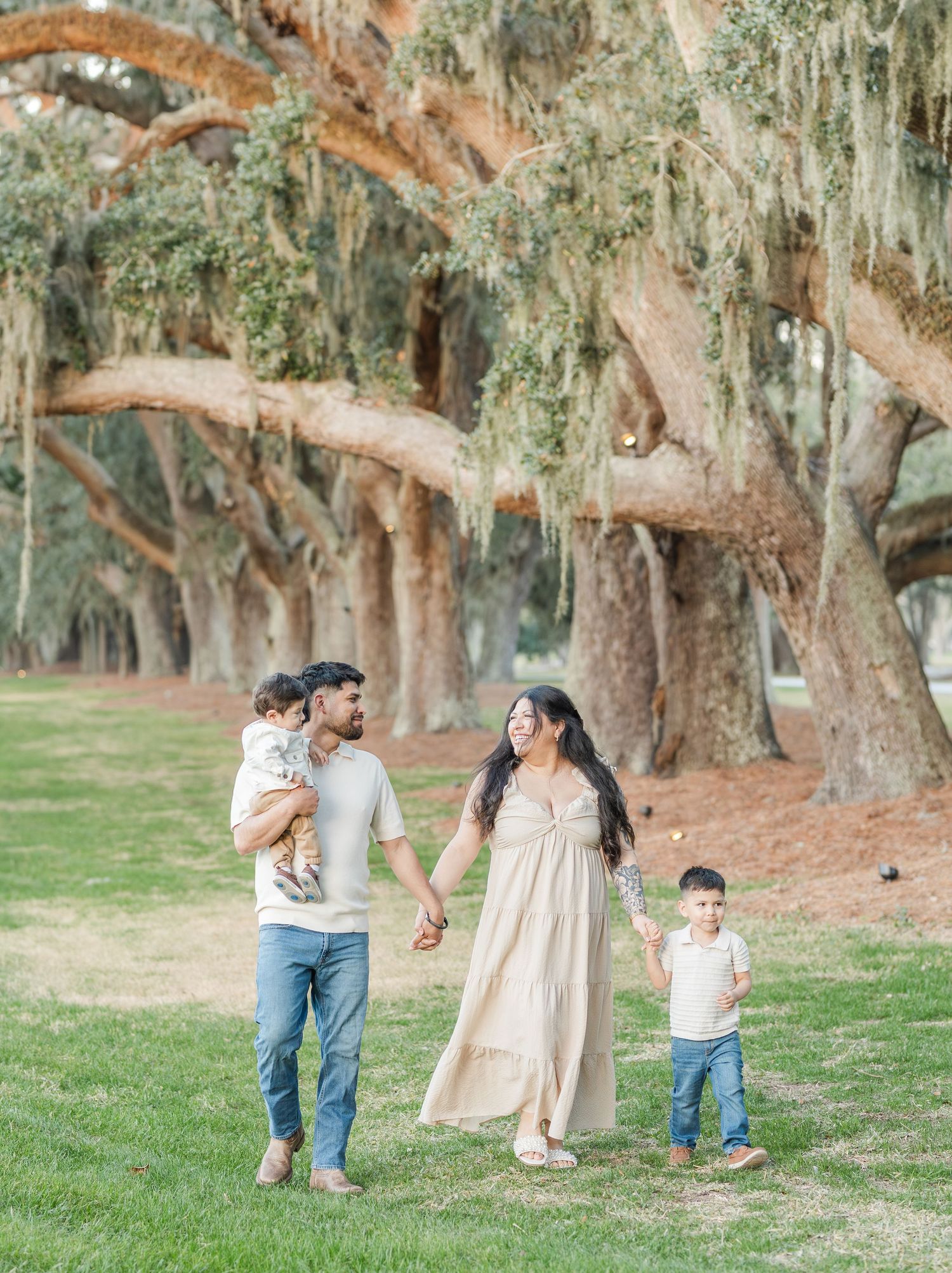 A family holds hands while strolling through a scenic park with moss-covered trees.
