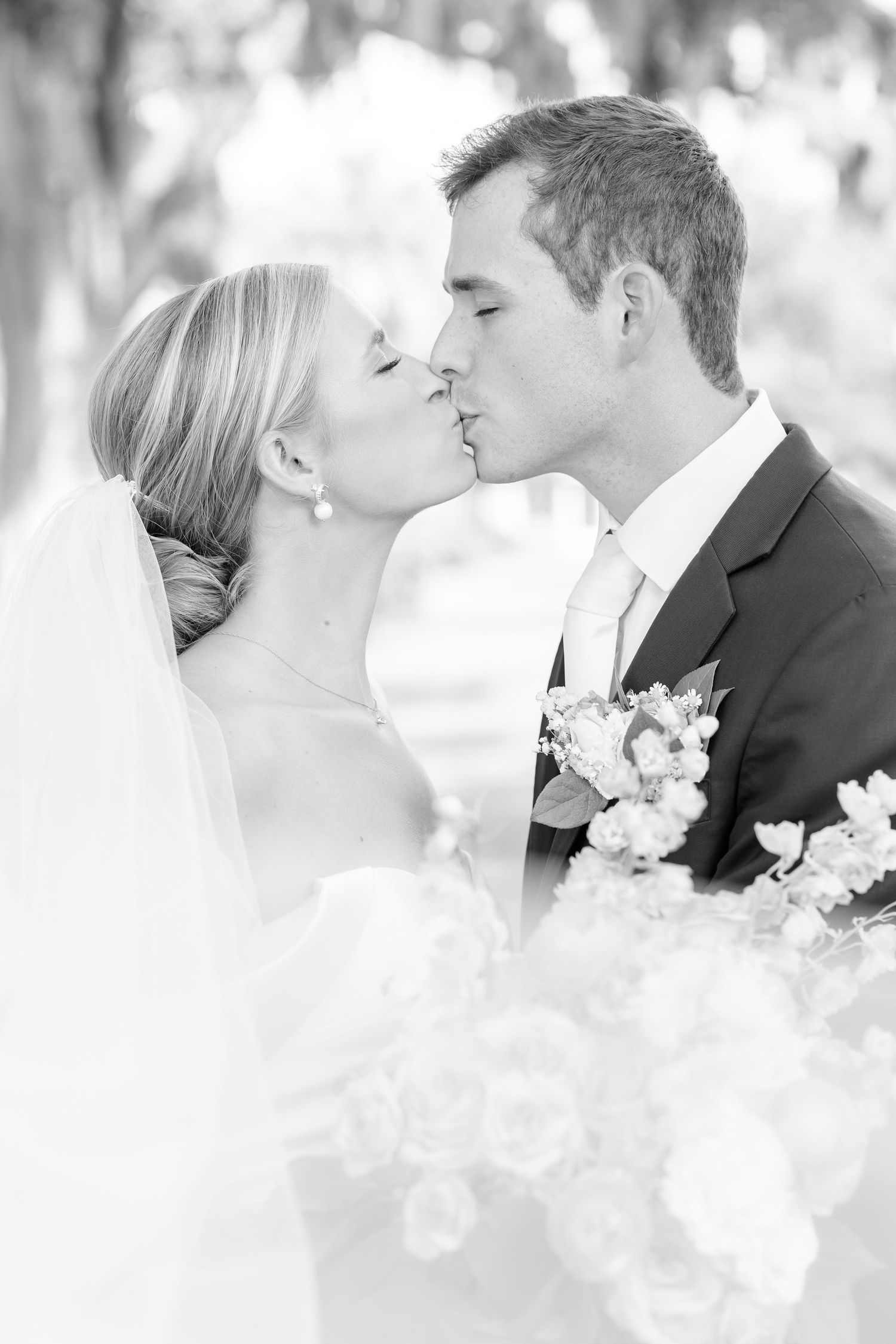 A romantic black and white wedding photo of a couple sharing a tender kiss with delicate flowers in the foreground.