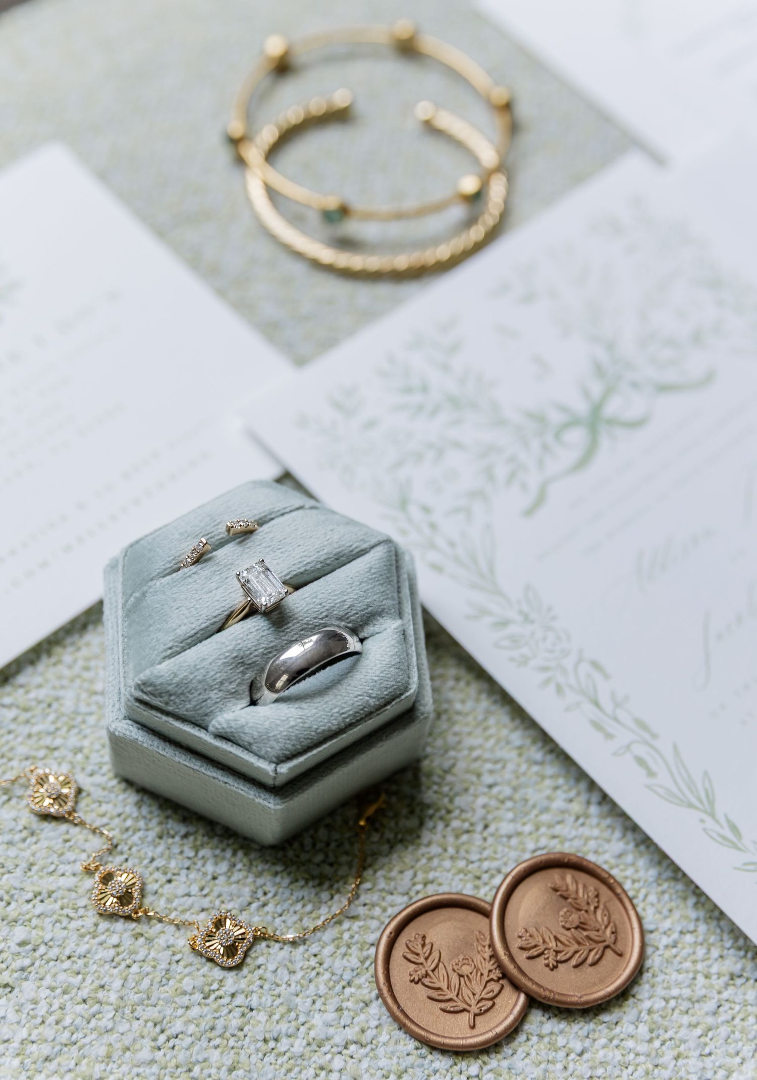 Wedding details featuring a sage green hexagonal ring box, gold bracelet, and wax seal stamps on delicate paper.