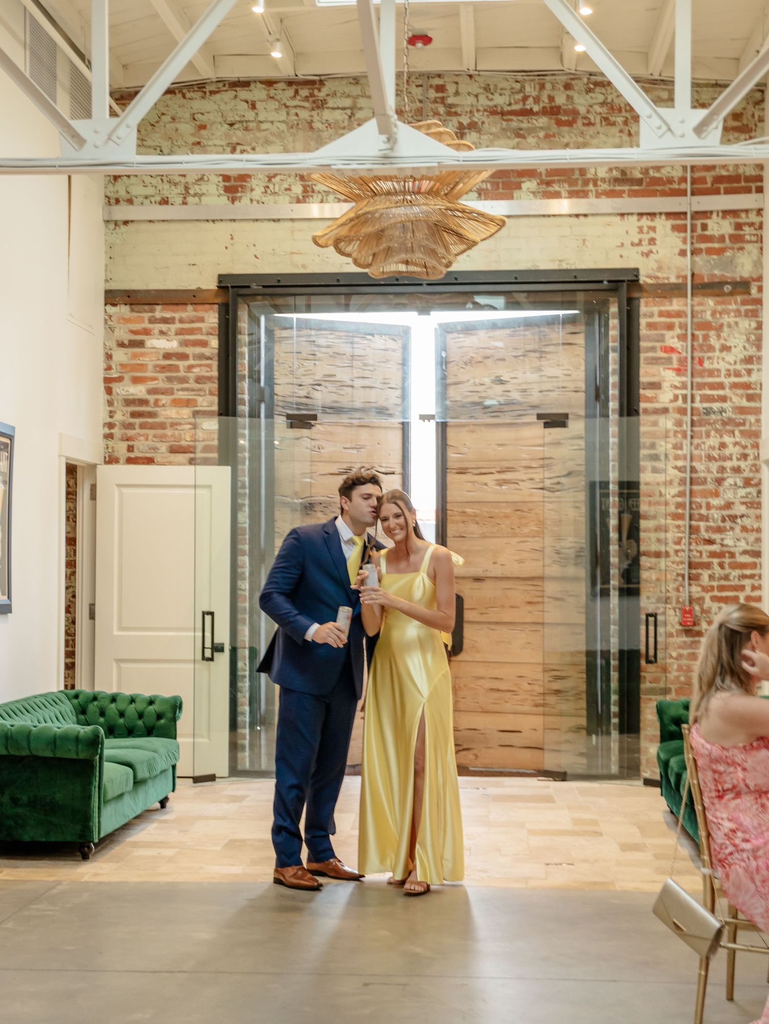 Couple in formal attire standing in elegant brick-walled venue with emerald green velvet couch and modern lighting.