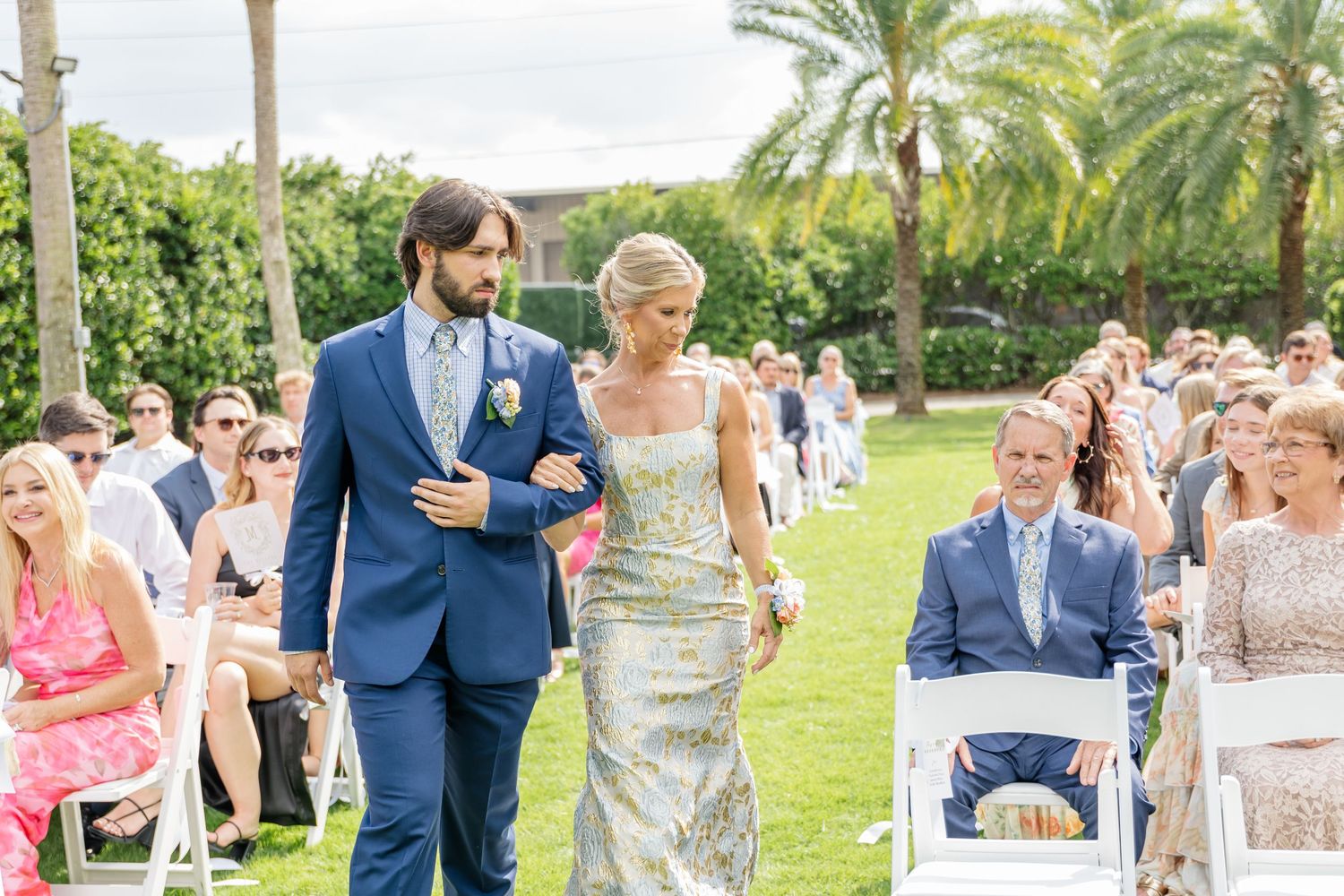 Wedding guests gather outdoors on a sunny day surrounded by palm trees and tropical landscaping.