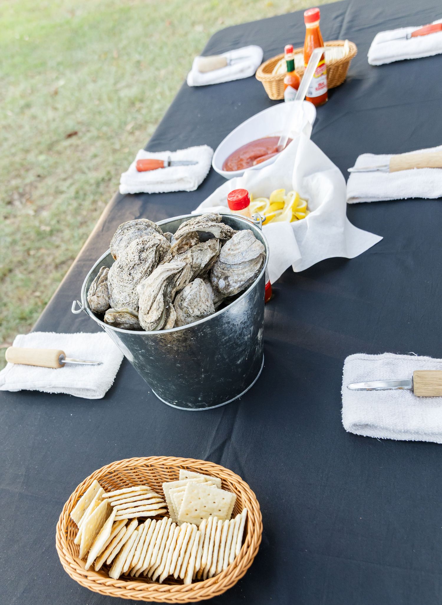 A rustic outdoor table setting with fresh oysters in a metal bucket and crackers in a woven basket on a black tablecloth.
