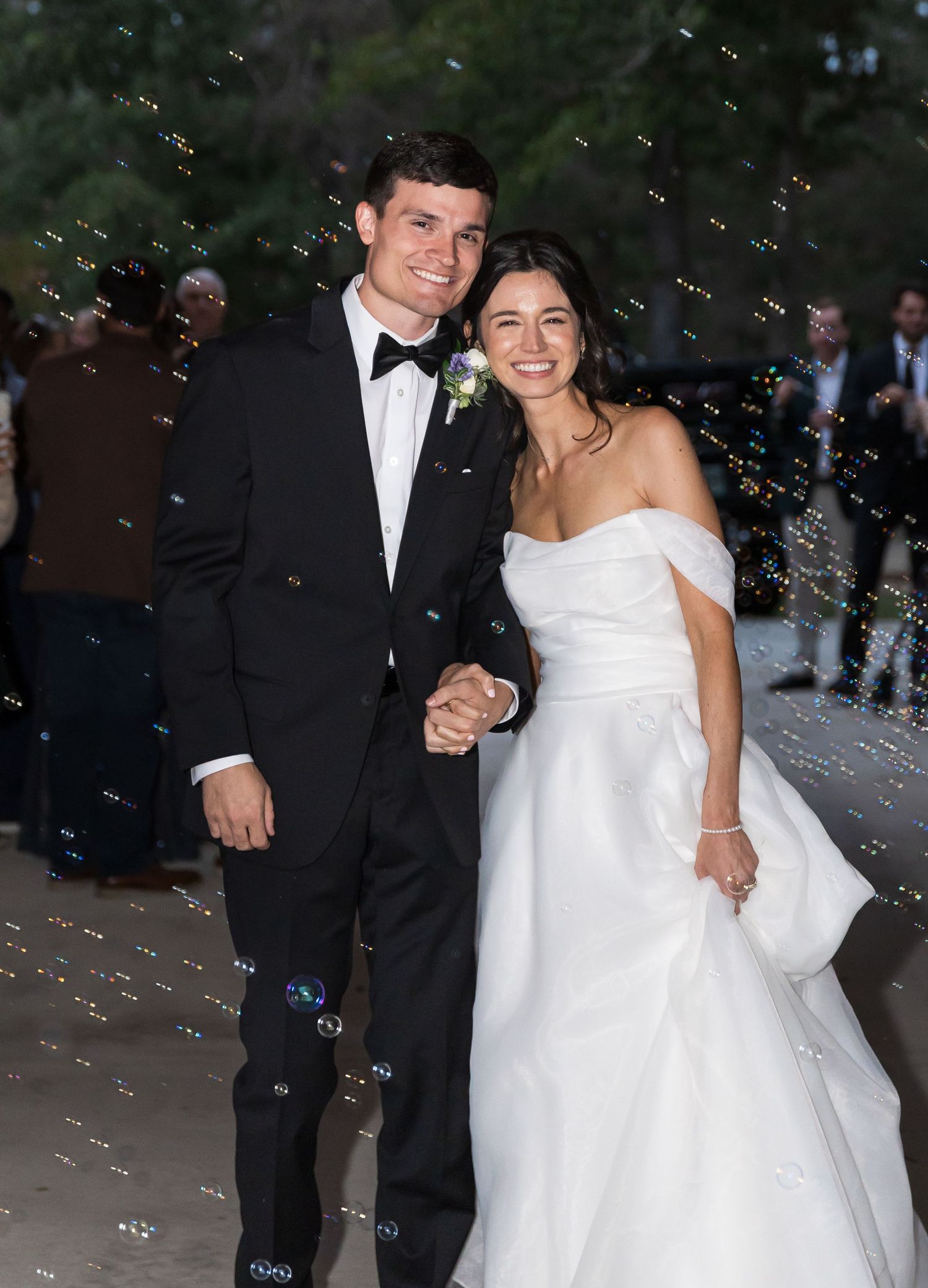 A couple celebrates their wedding day outdoors at dusk with floating bubbles creating a magical atmosphere.