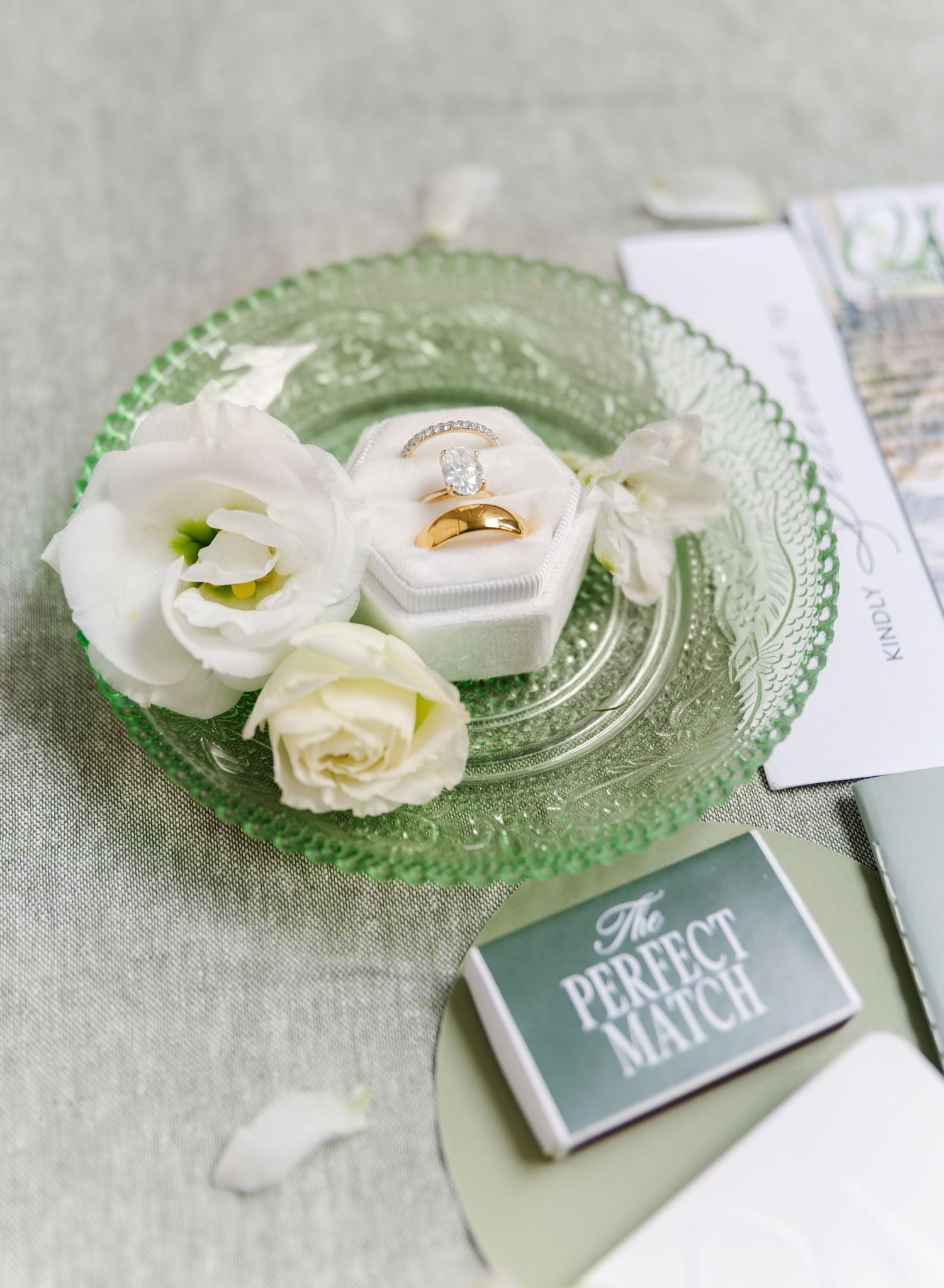 Wedding rings displayed on green glass plate with white roses and 'The Perfect Match' card.