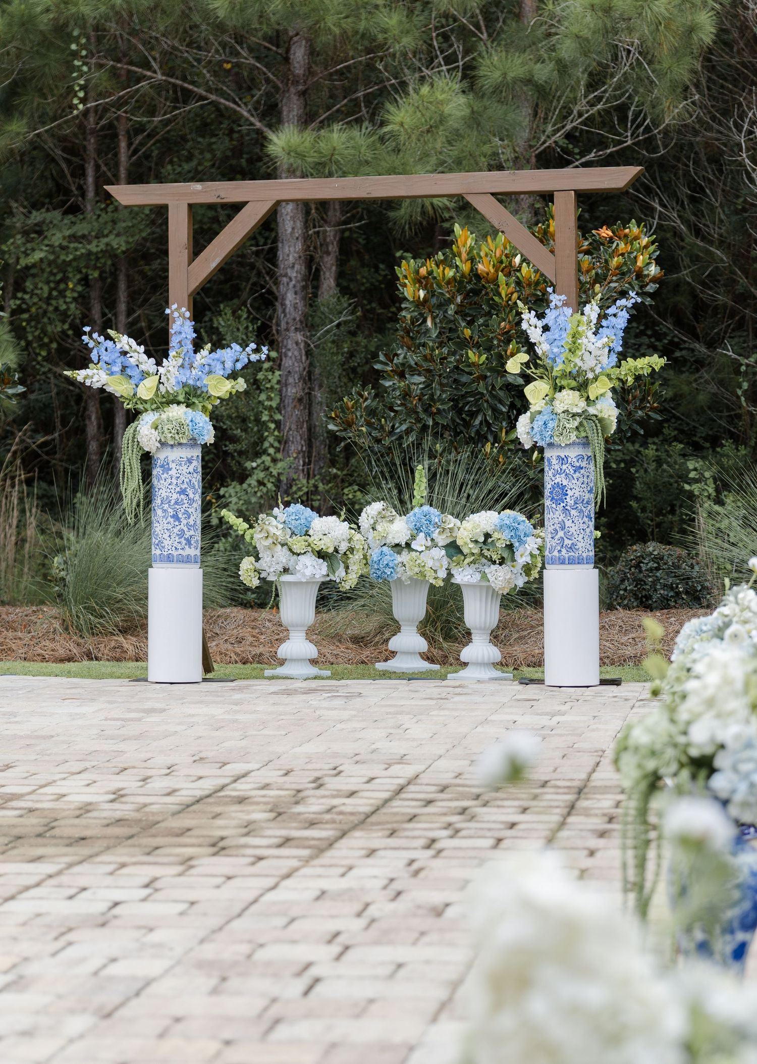 Elegant outdoor wedding ceremony arch with blue and white floral arrangements on white pedestals against a forest backdrop.