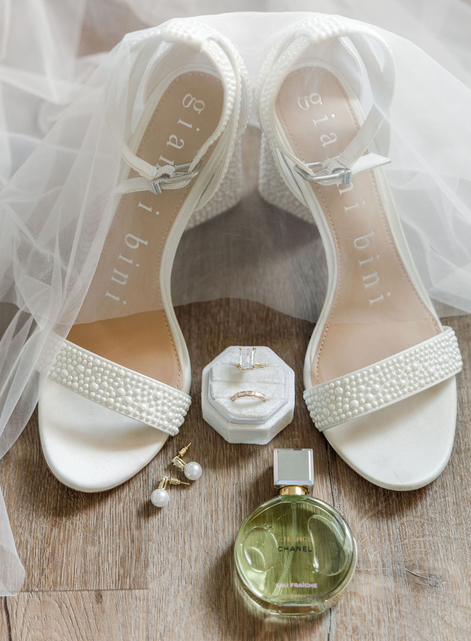 Pearl-embellished bridal heels by Gianni Bini displayed with Chanel perfume, gold ring, and pearl earrings on wood floor.
