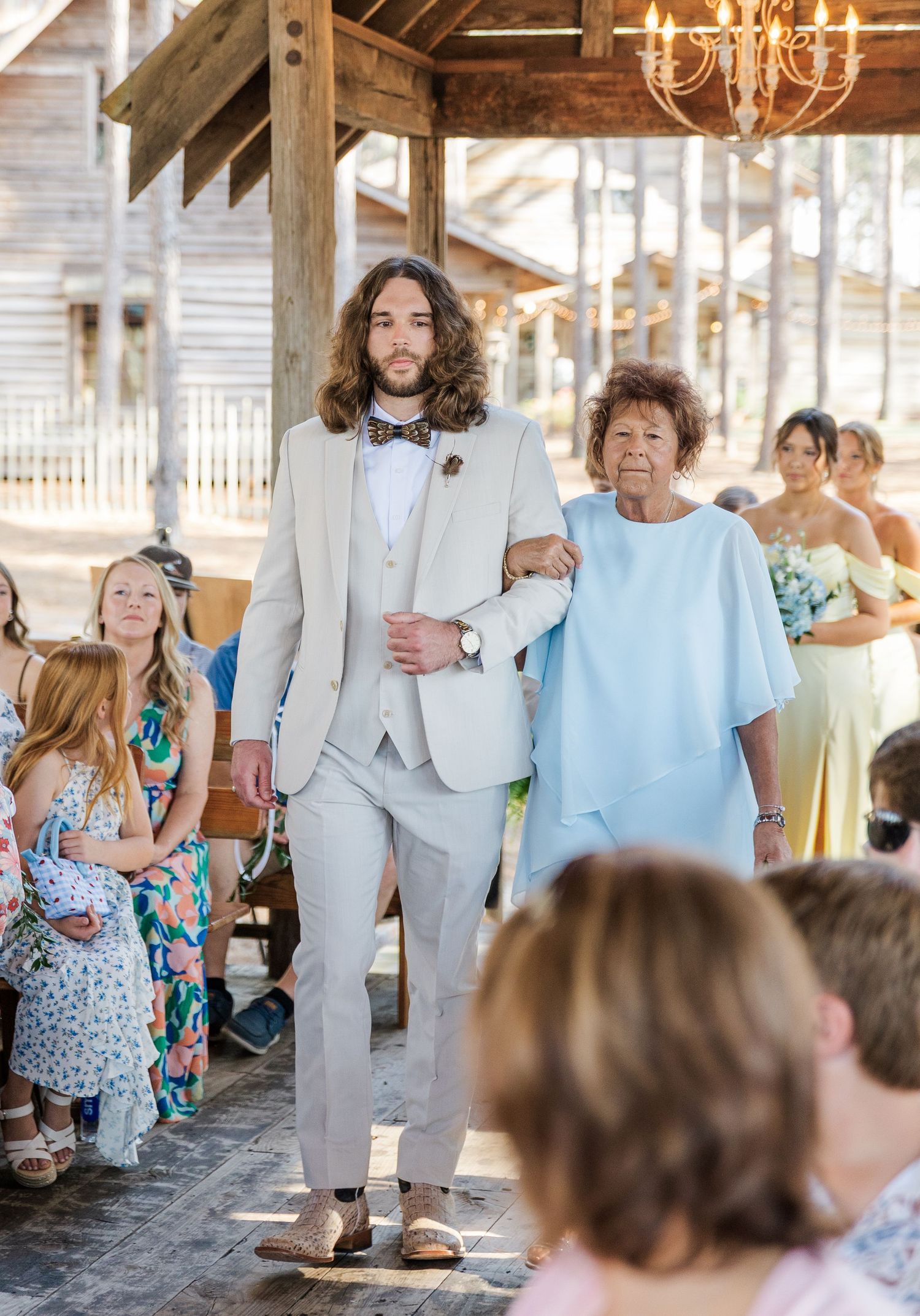 Groomsman in tan three-piece suit escorts elderly woman down aisle at rustic outdoor wedding ceremony.
