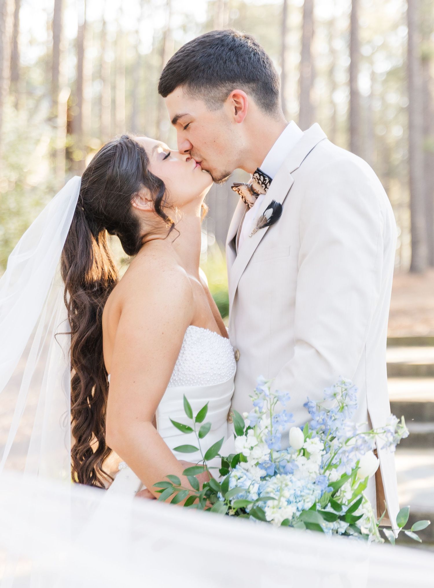 Bride and groom share a romantic kiss outdoors, with blue floral bouquet and pine trees in background.