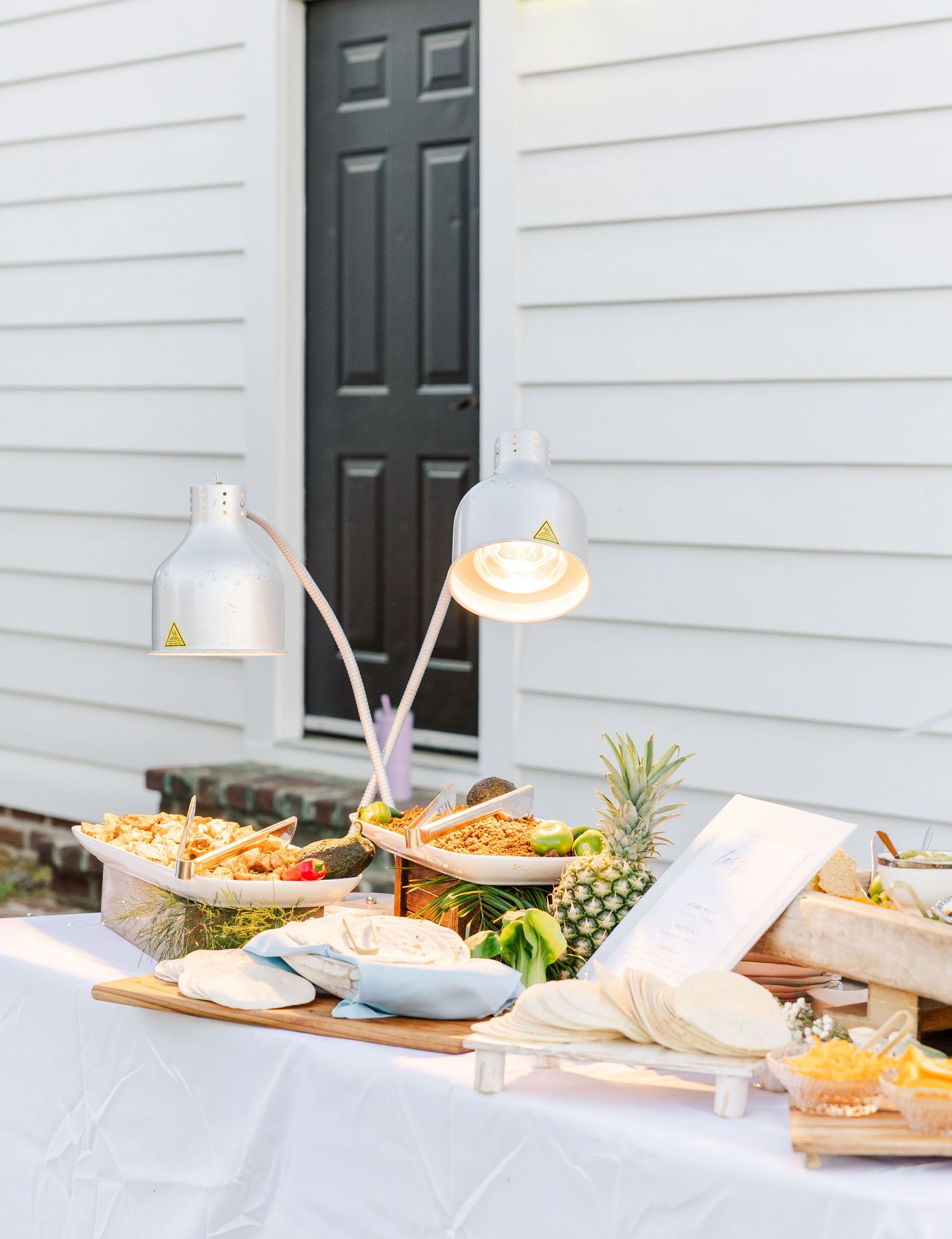 Outdoor wedding food station with warming lamps, tropical fruits, and assorted dishes.