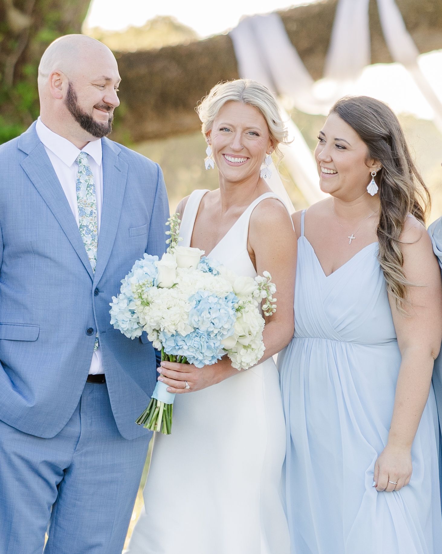 Smiling bride in white gown holding blue and white hydrangea bouquet flanked by wedding party members.