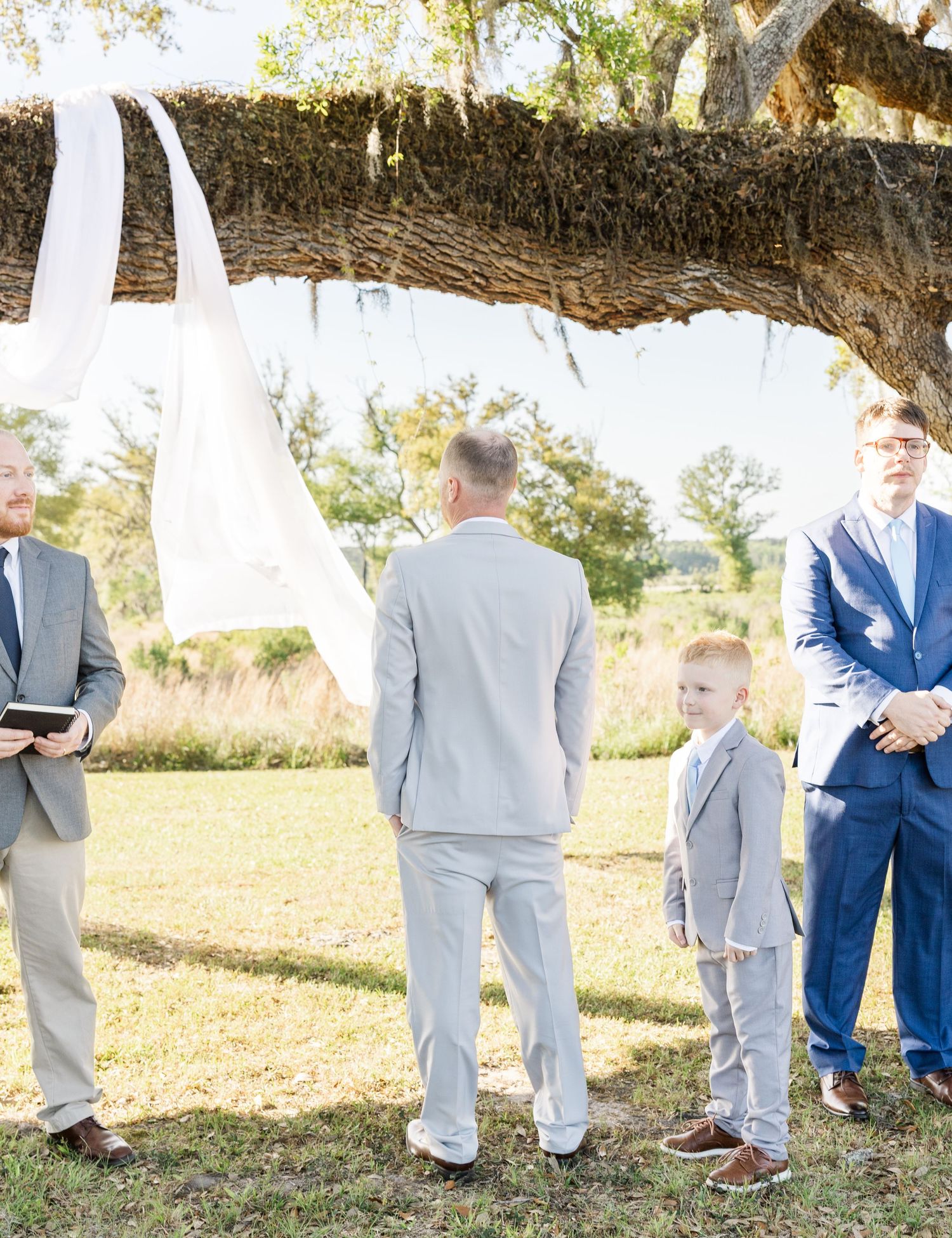 Outdoor wedding ceremony under a large oak tree with grooms and young ring bearer in matching gray suits.