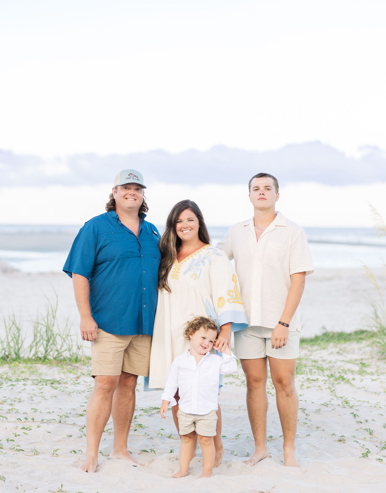 Family group in blue and white casual attire poses together for beach photos at sunset.