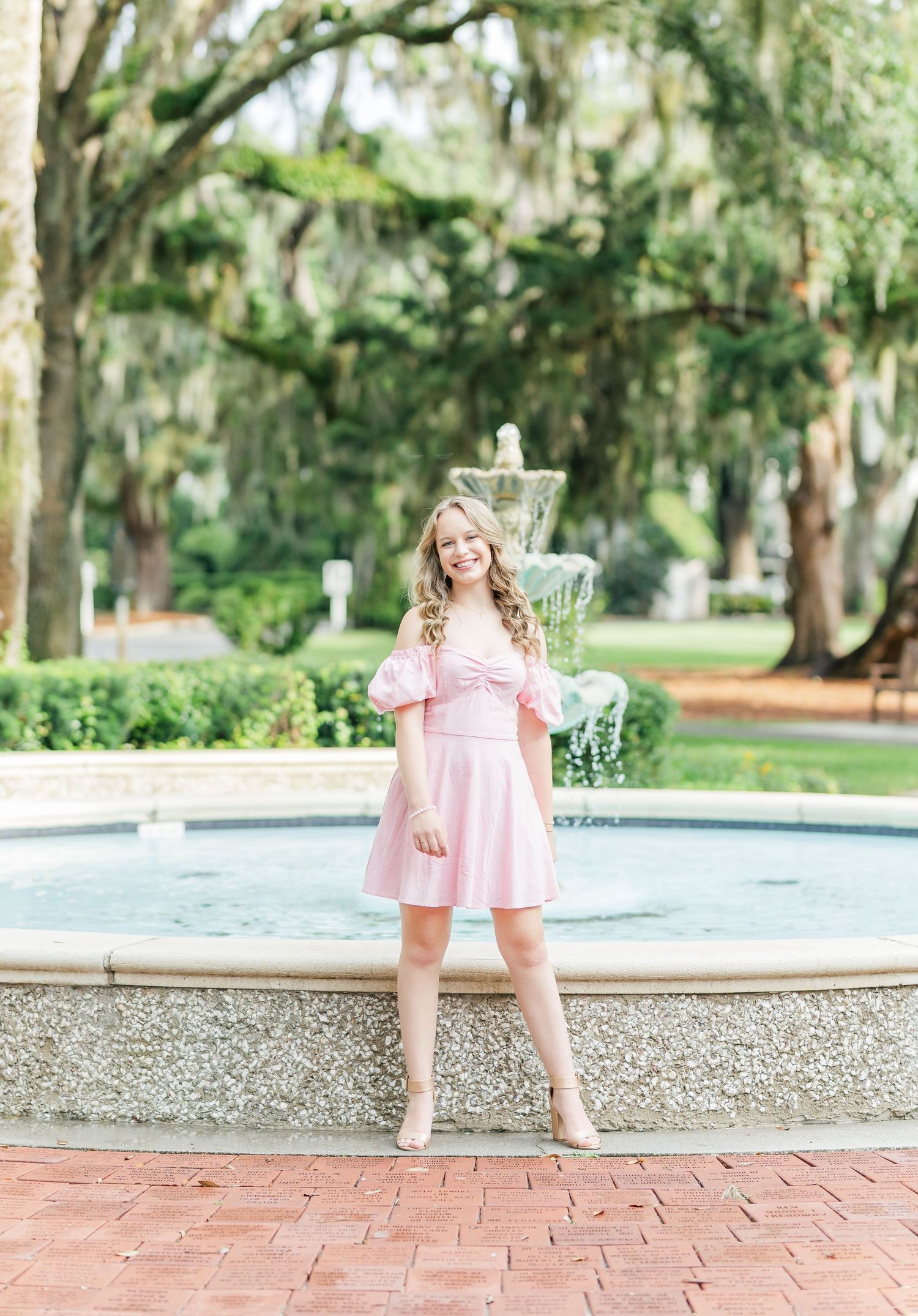 A person in a pale pink dress stands elegantly beside a garden fountain with Spanish moss trees in background.