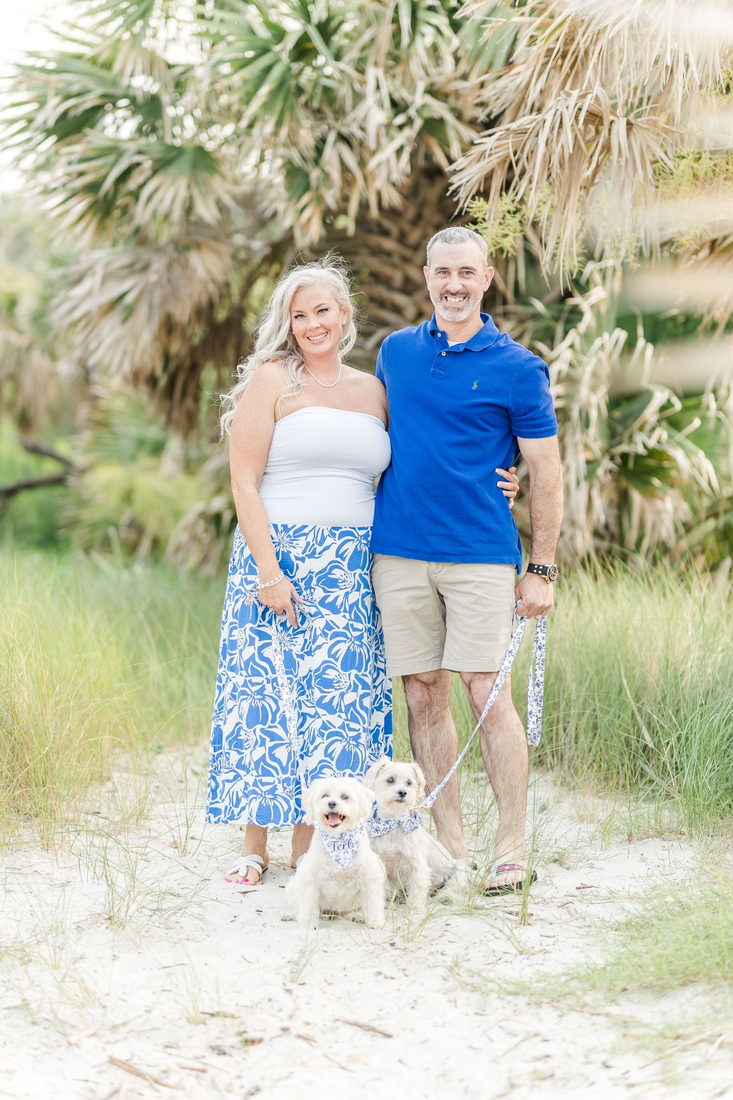 A couple poses with their small white dog on a palm-tree lined beach path.