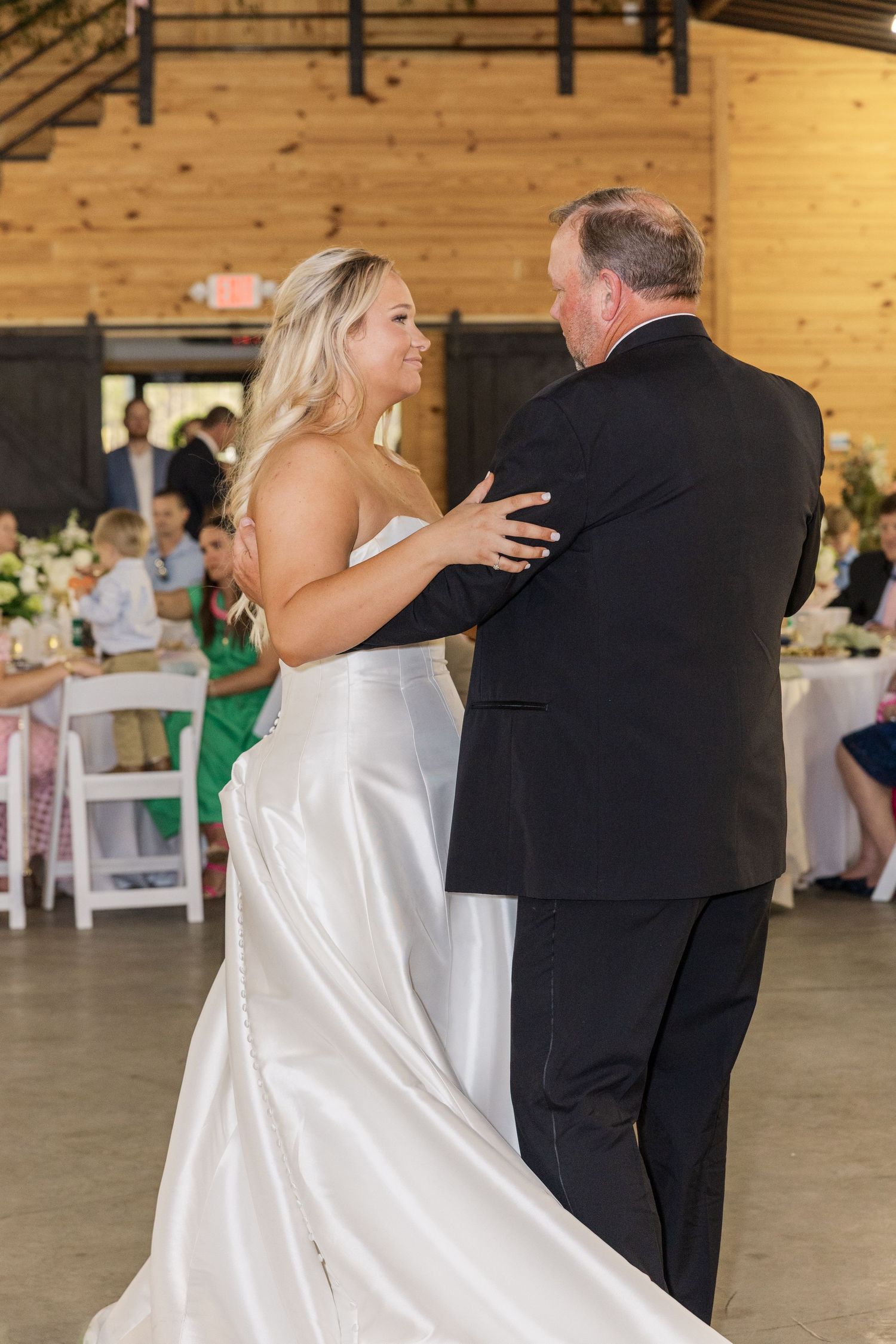 Bride in white satin gown shares father-daughter dance at rustic barn wedding reception.