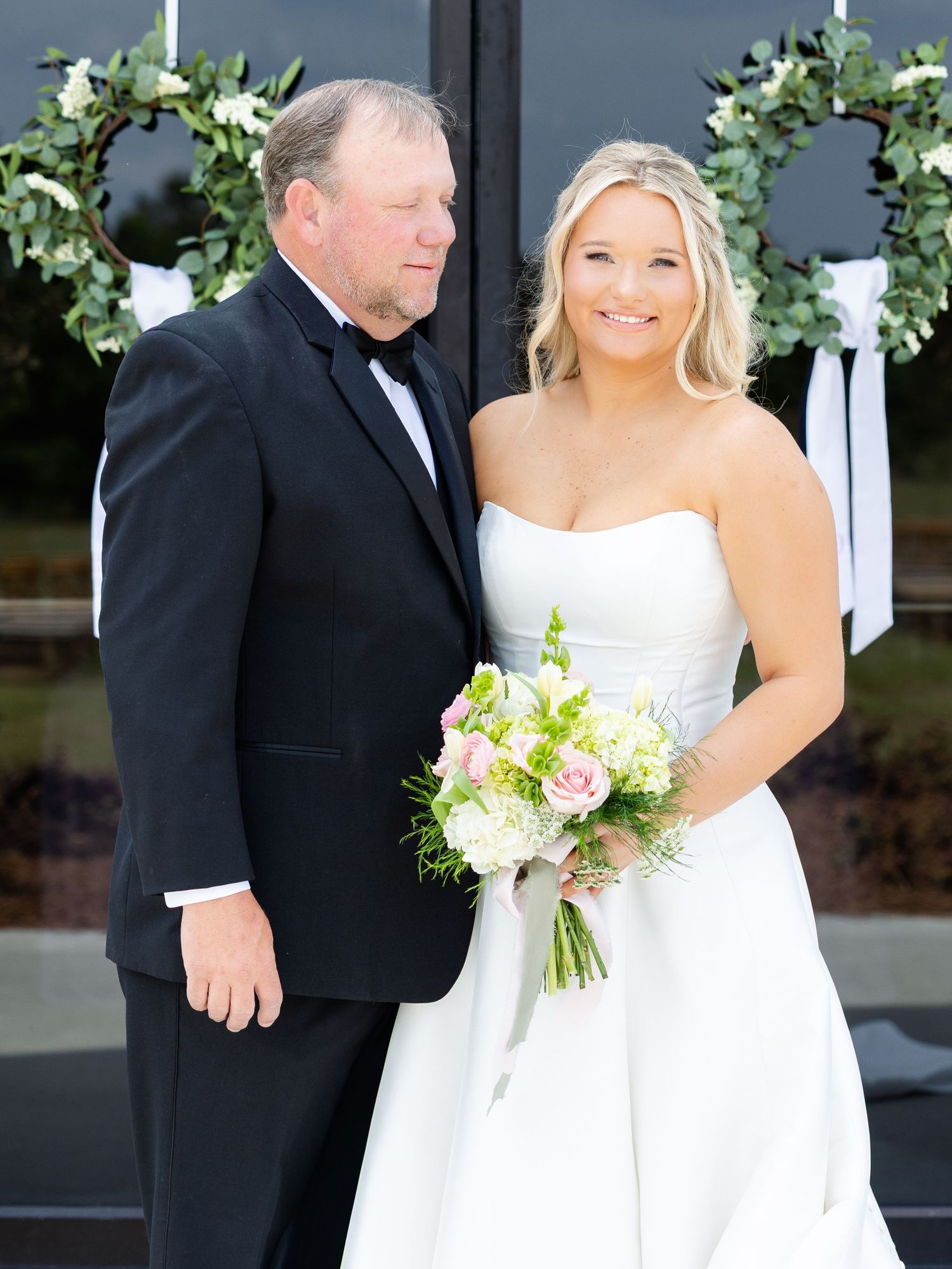Blonde bride in strapless white gown holding pink bouquet poses with father in black tuxedo.