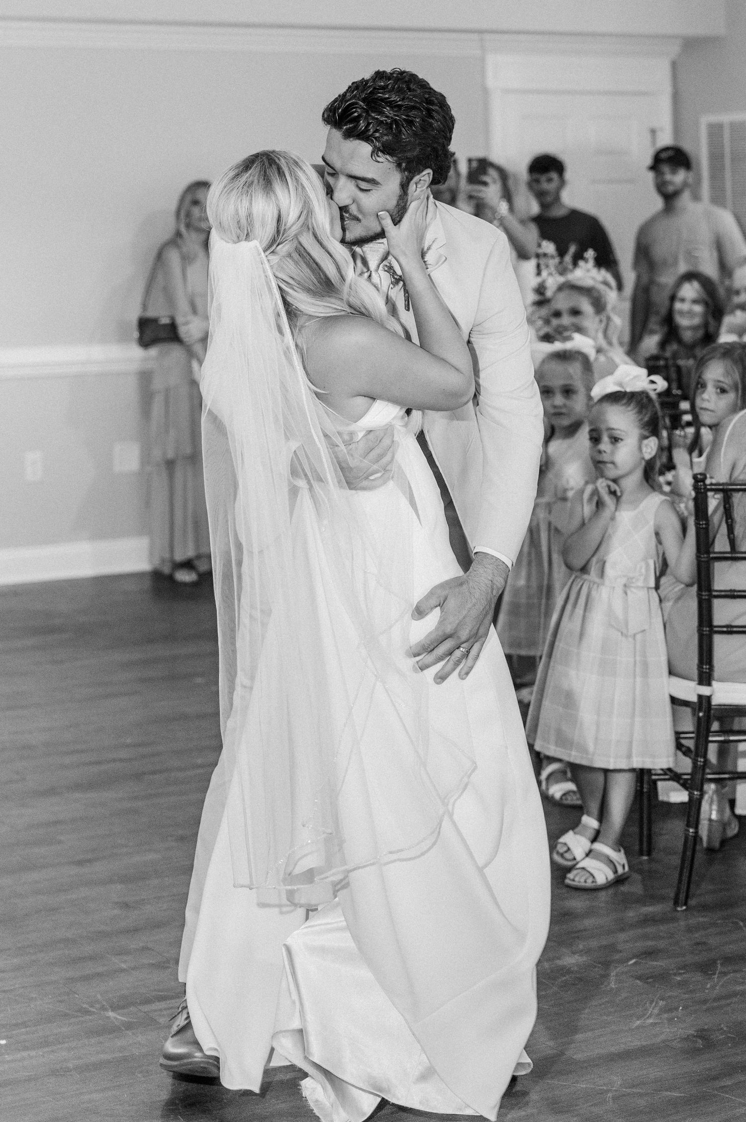 Bride and groom share their first dance kiss while flower girls watch at elegant wedding reception.