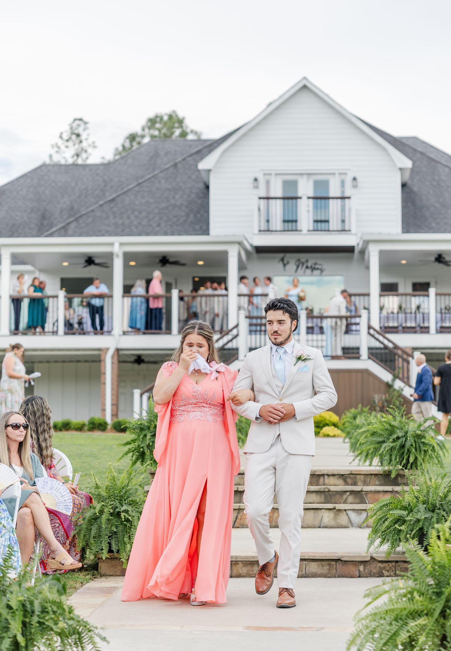Groom in beige suit escorts a woman in coral dress down the aisle at an outdoor wedding ceremony.