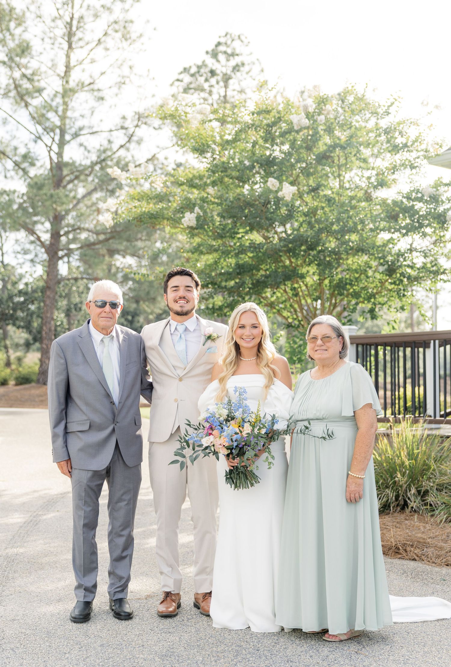 Bride and groom pose with parents outdoors, bride holding a blue and white bouquet on a sunny wedding day.