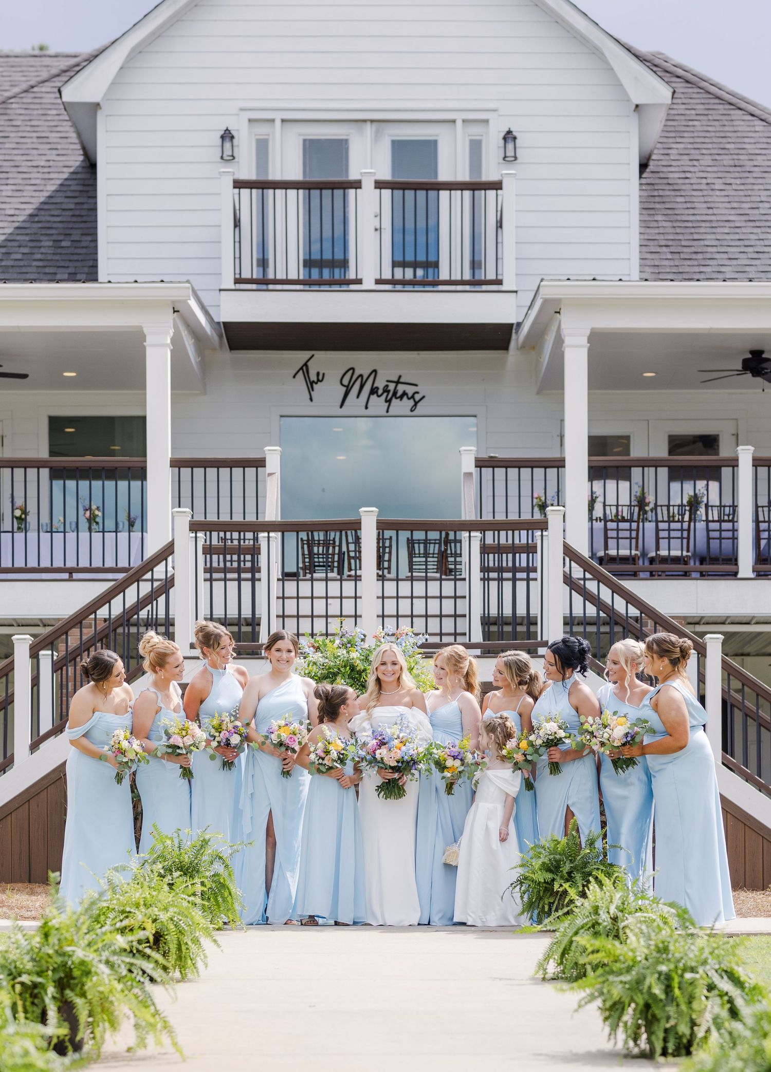 Bride in white gown poses with bridesmaids in light blue dresses holding colorful bouquets before a white venue.
