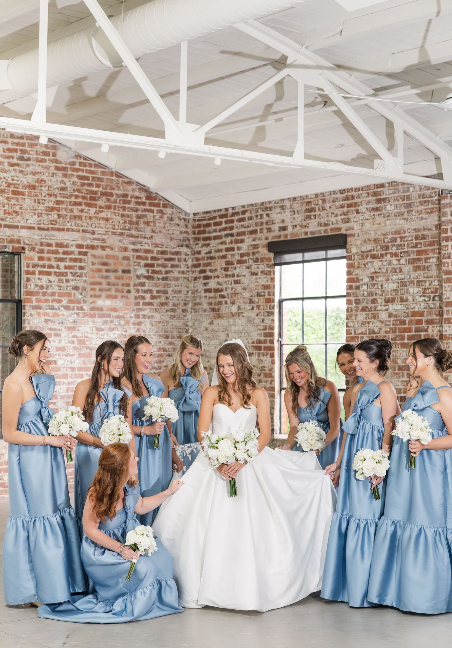 Bride in white gown surrounded by bridesmaids in dusty blue dresses holding white bouquets in rustic brick venue.