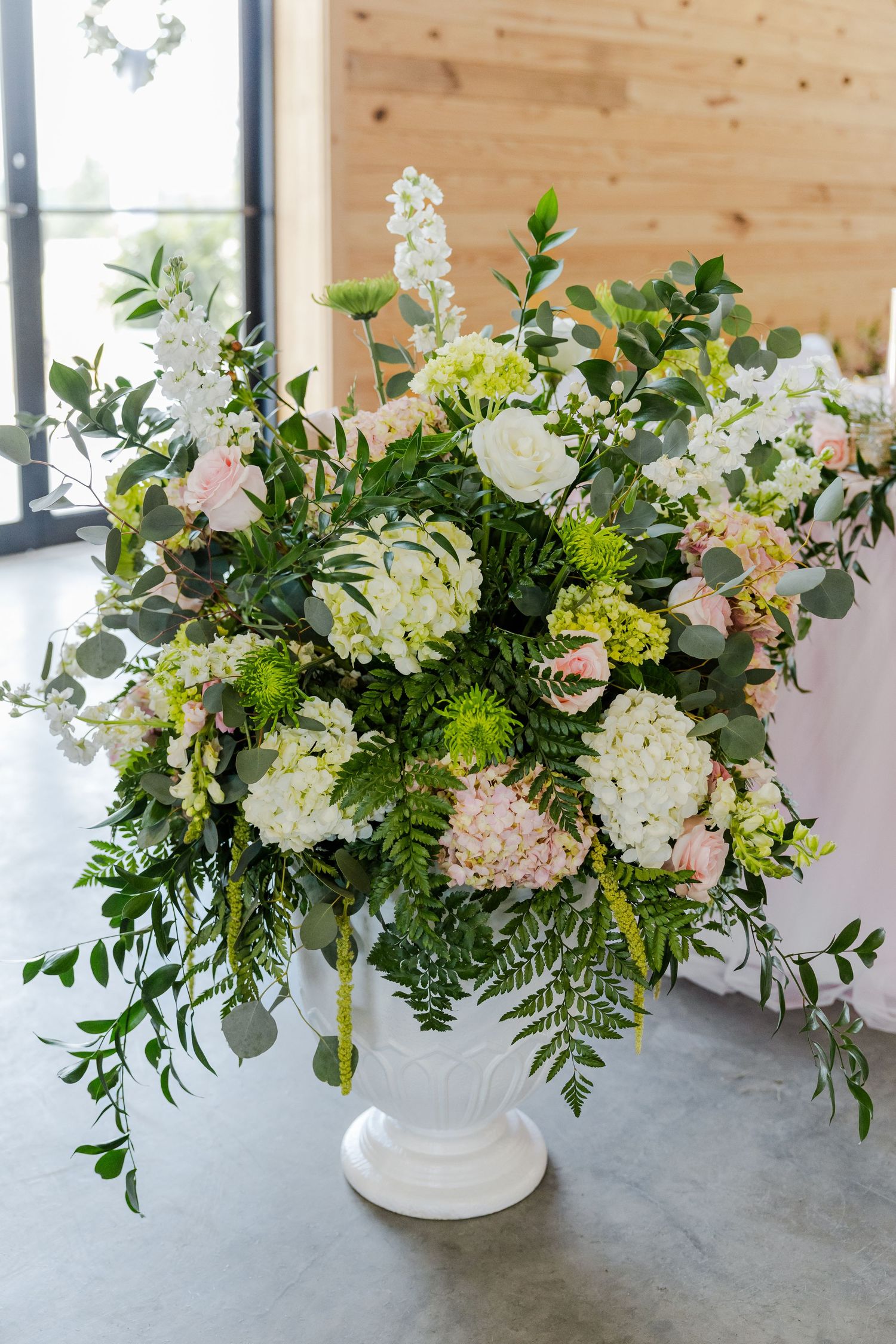 Large white urn vase with lush wedding floral arrangement featuring white hydrangeas, pink roses, and greenery.