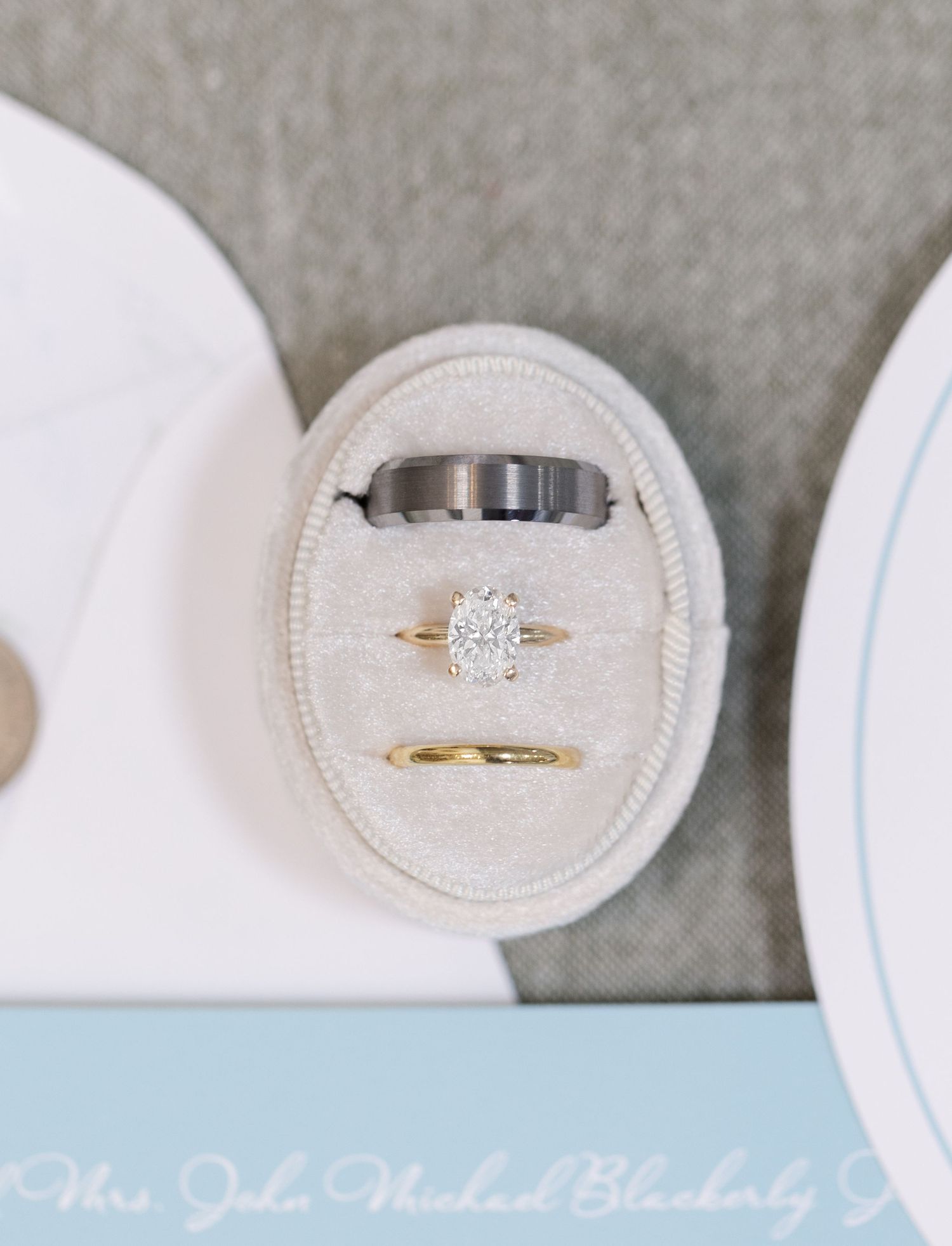 Three wedding rings displayed in an oval velvet ring box, featuring a diamond engagement ring, gold band, and tungsten band.