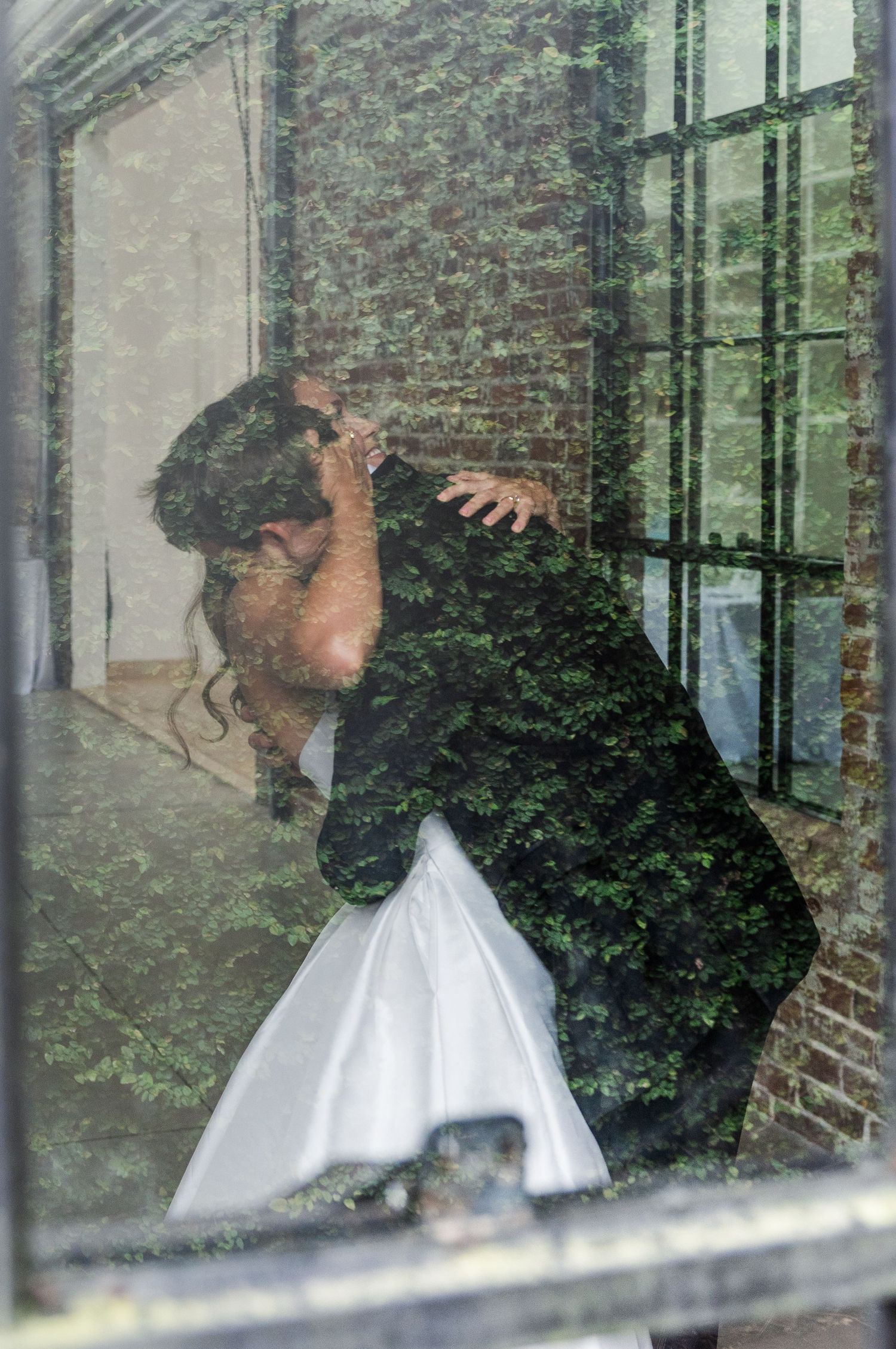 Bride and groom sharing first look kiss captured through window with lush green foliage reflection.
