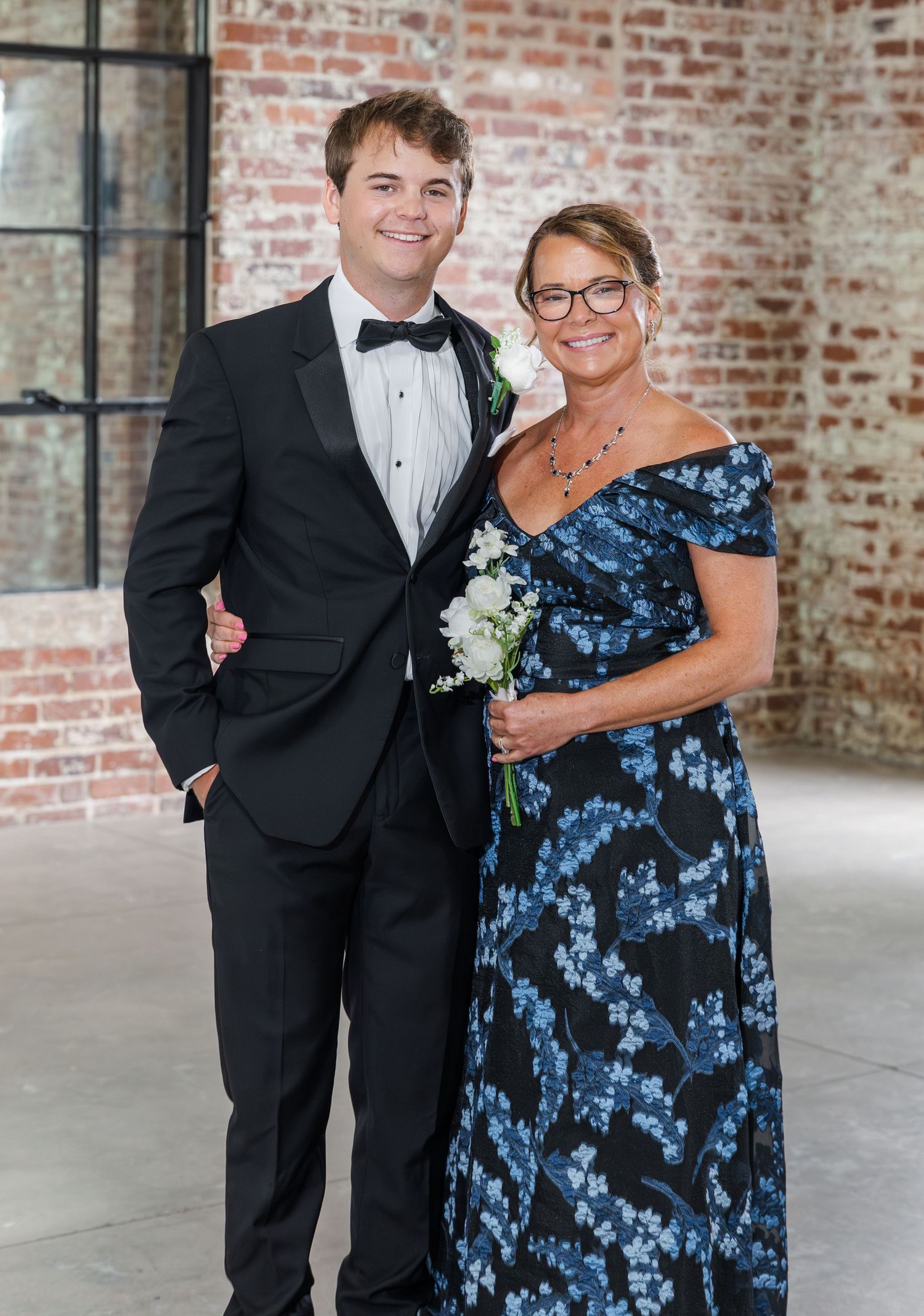 Groom in black tuxedo poses with mother in blue floral off-shoulder gown at wedding venue.