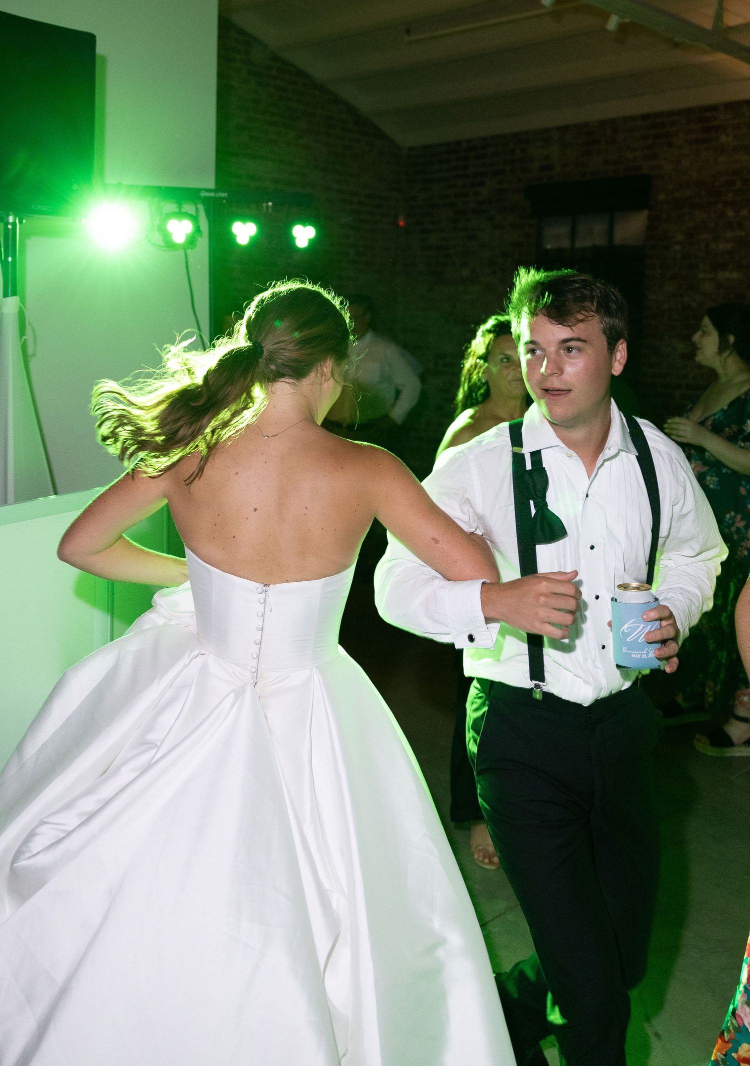 Bride in white gown dances with groomsman in suspenders under green lights at wedding reception.