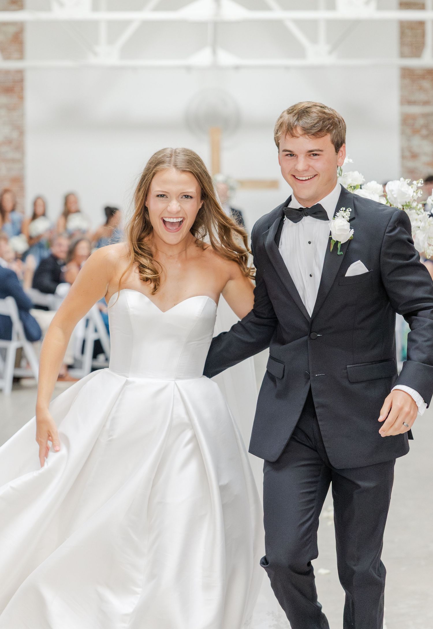 Joyful bride in white strapless gown and groom in black tuxedo recess after wedding ceremony.