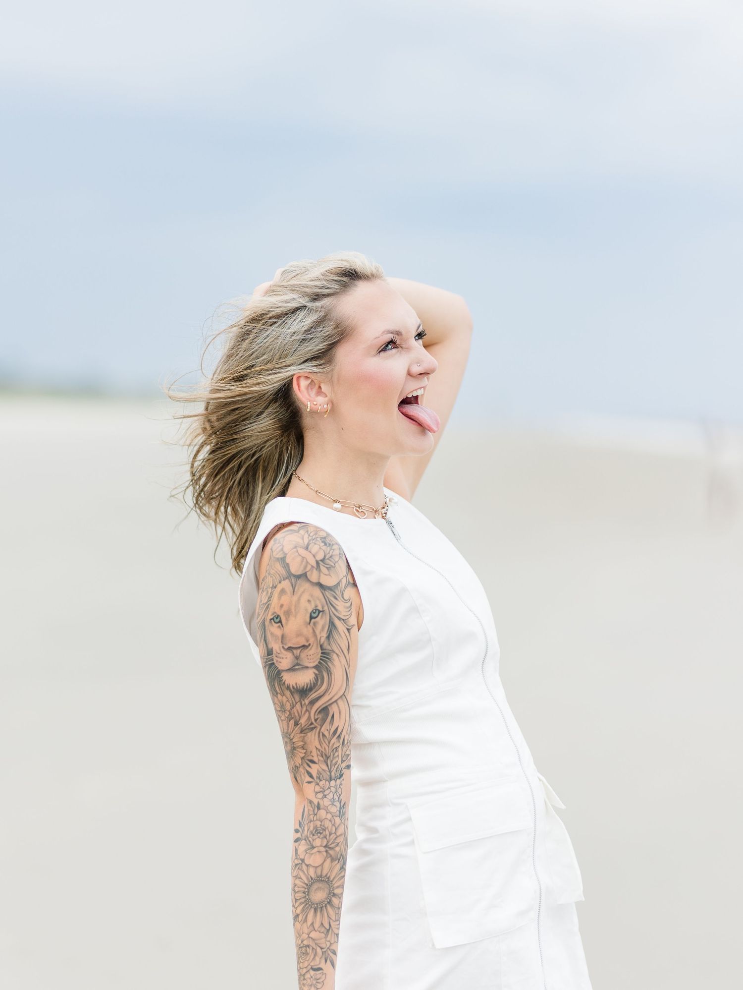 Playful beach portrait featuring white dress and artistic tattoo sleeve against cloudy skies.