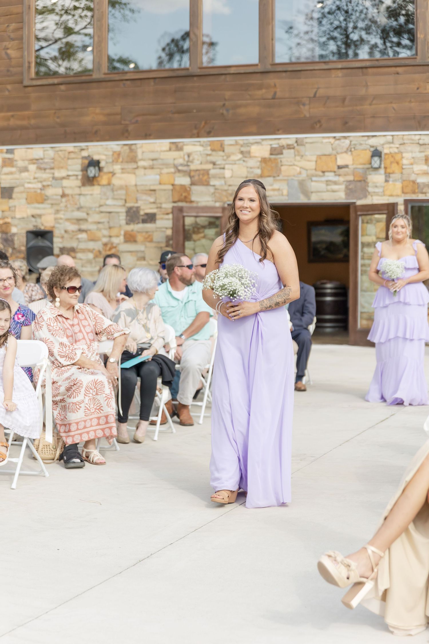 Bridesmaid carrying bouquet walks processional at outdoor stone venue.