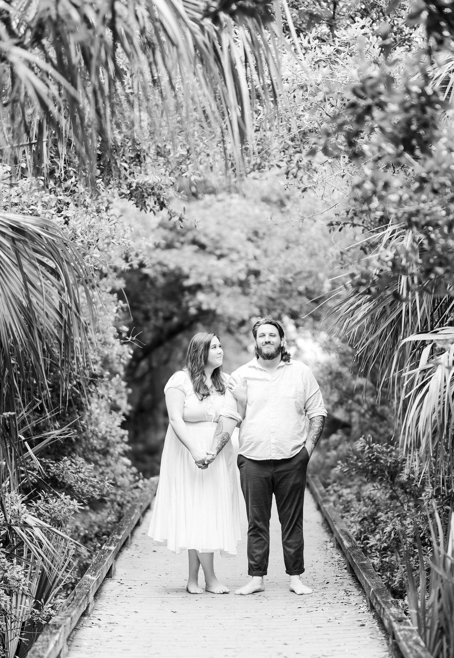 A romantic couple stands together on a garden path framed by tropical palm fronds and lush greenery.