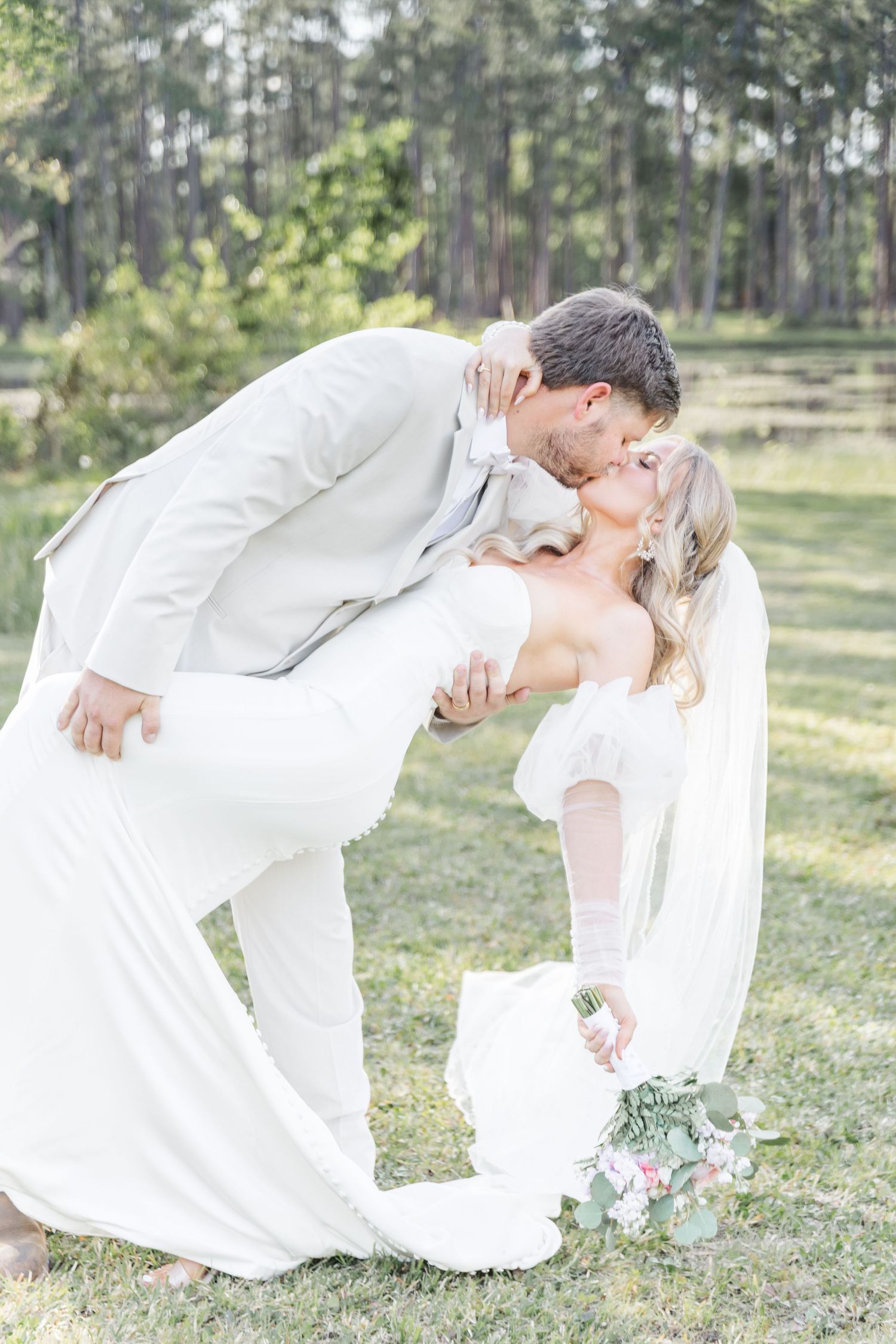 A romantic wedding couple shares a dramatic dip kiss outdoors with pine trees in the background.