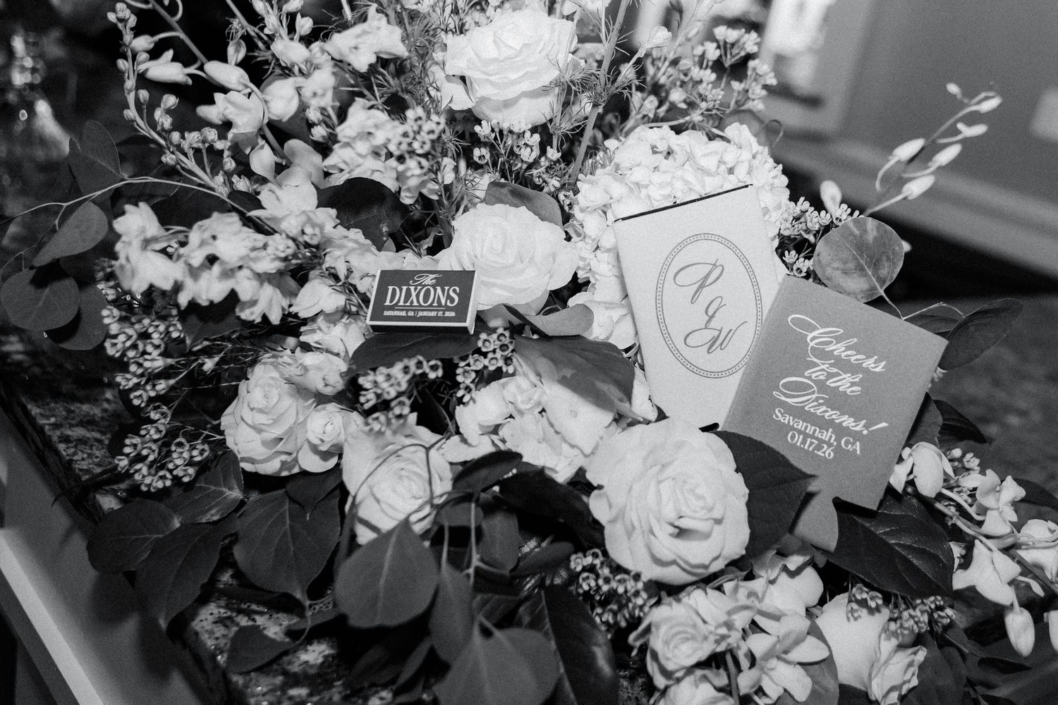 Wedding details featuring white roses and invitation cards arranged on a dark surface in black and white.