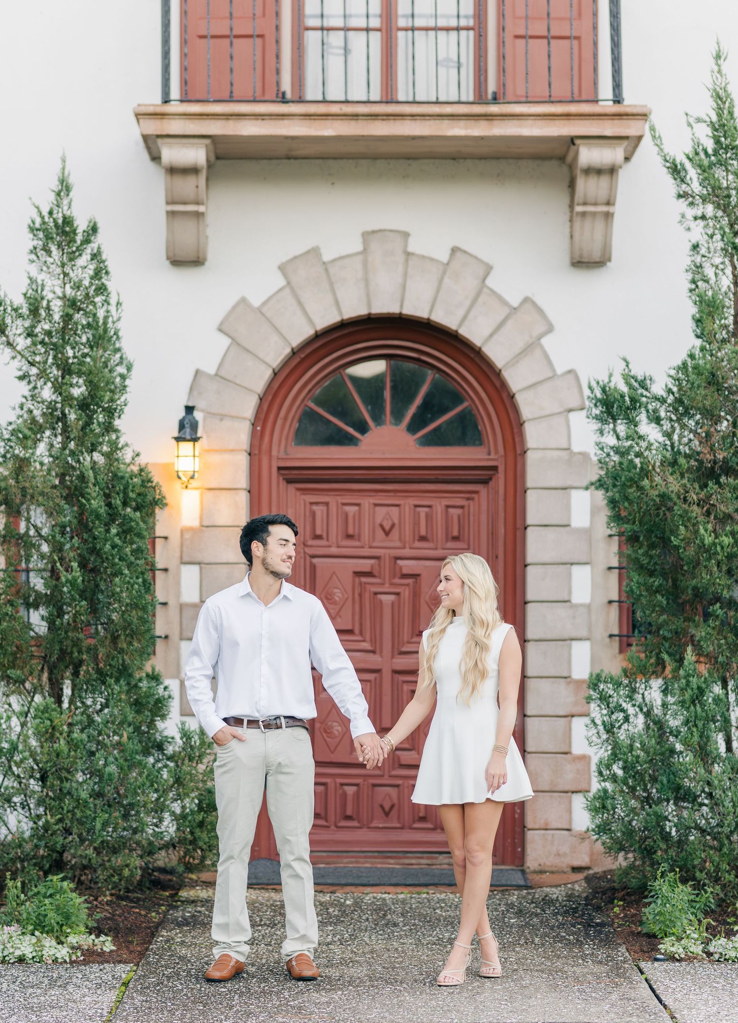 Romantic couple in white outfits posing against a building with red doors and archways.