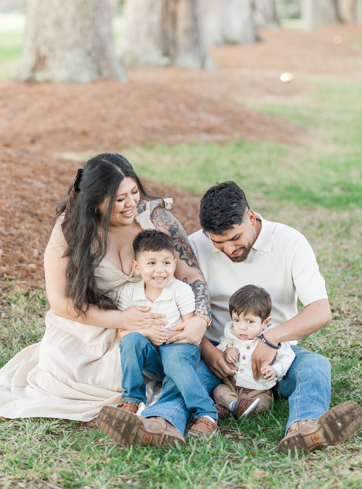 A family sits together on fallen leaves beneath towering trees in a park setting.