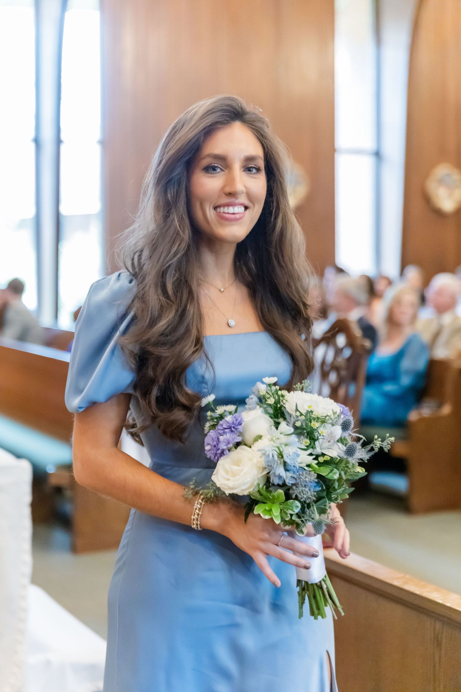 Bridesmaid in light blue dress holding a bouquet of white and purple flowers in church.